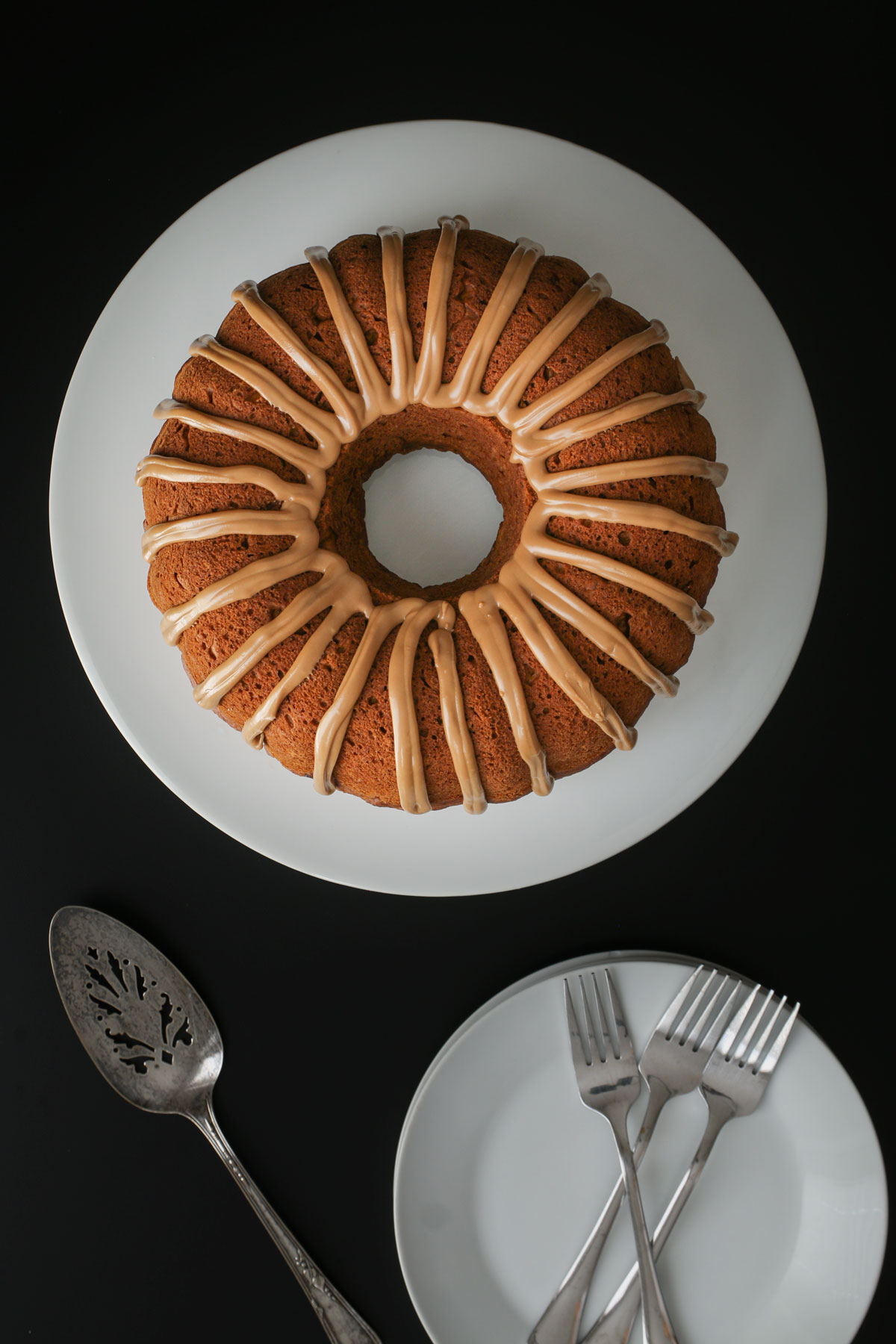overhead shot of pumpkin bundt cake on white platter with cake server on black table next to stack of plates with forks.