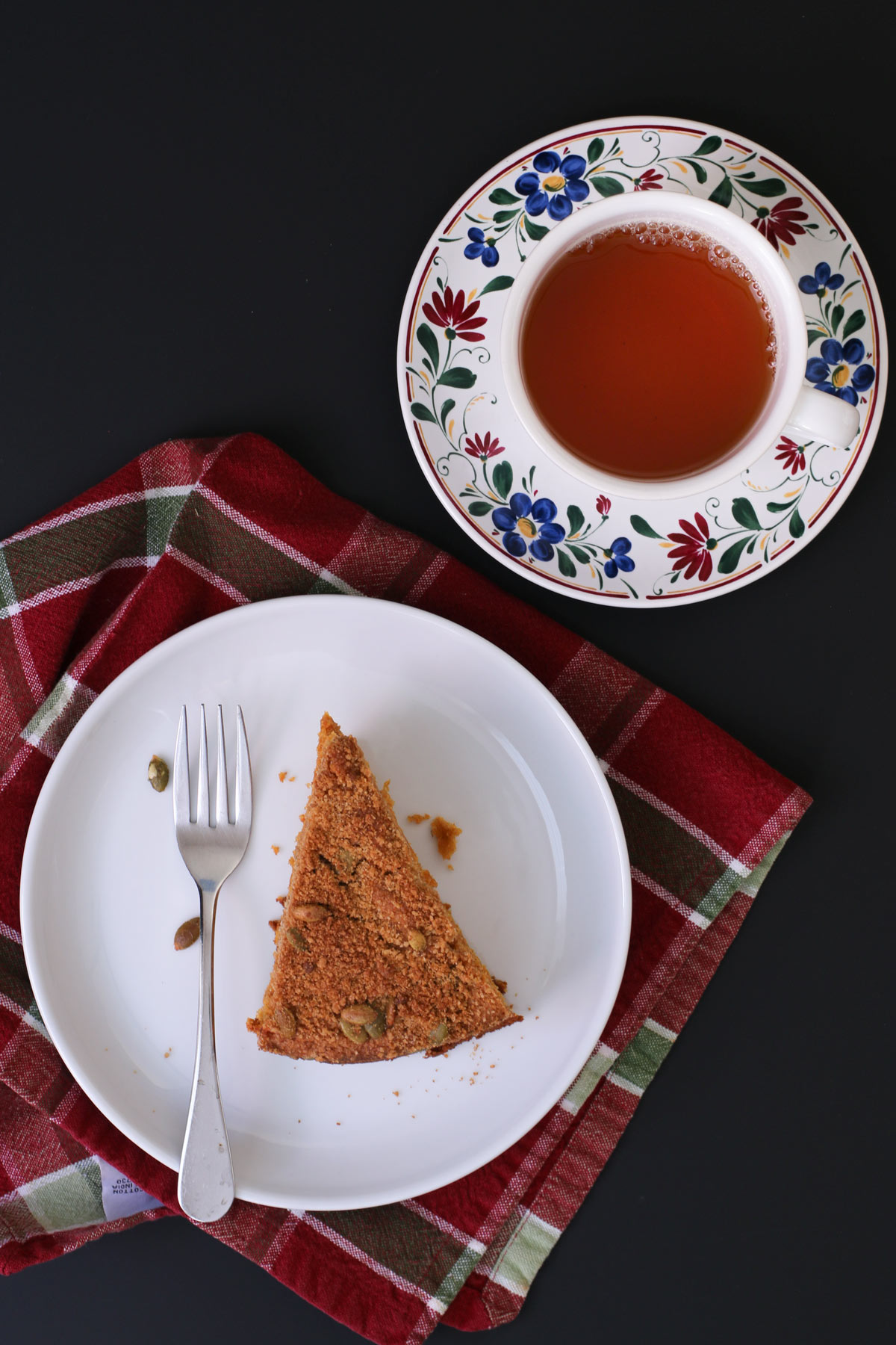 wedge of pumpkin coffee cake on white plate atop red and green plaid napkin with a cup of tea on a flowered saucer nearby.