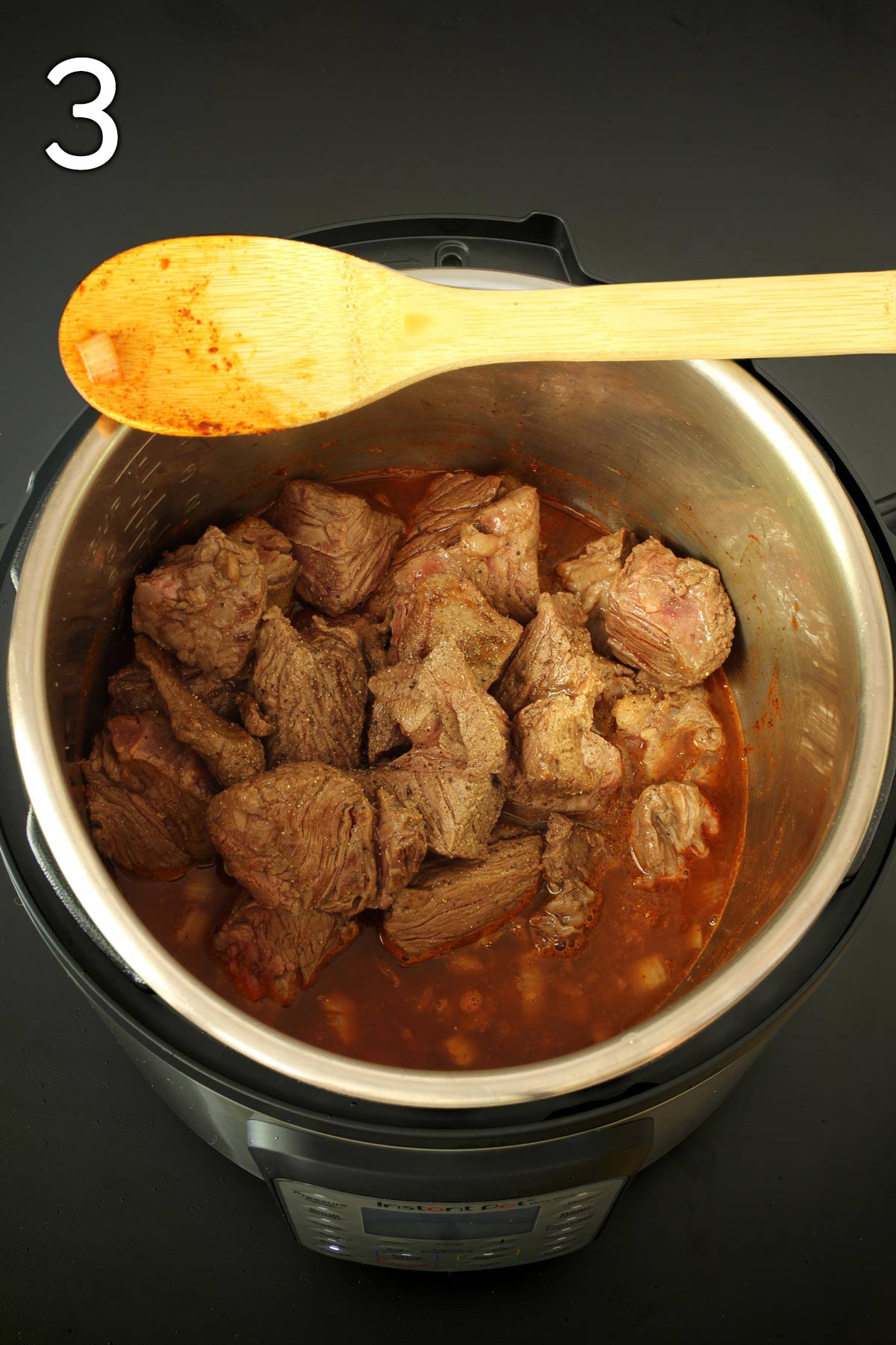 beef cubes back in the pot with sauce, wooden spoon resting across the top of the pot.