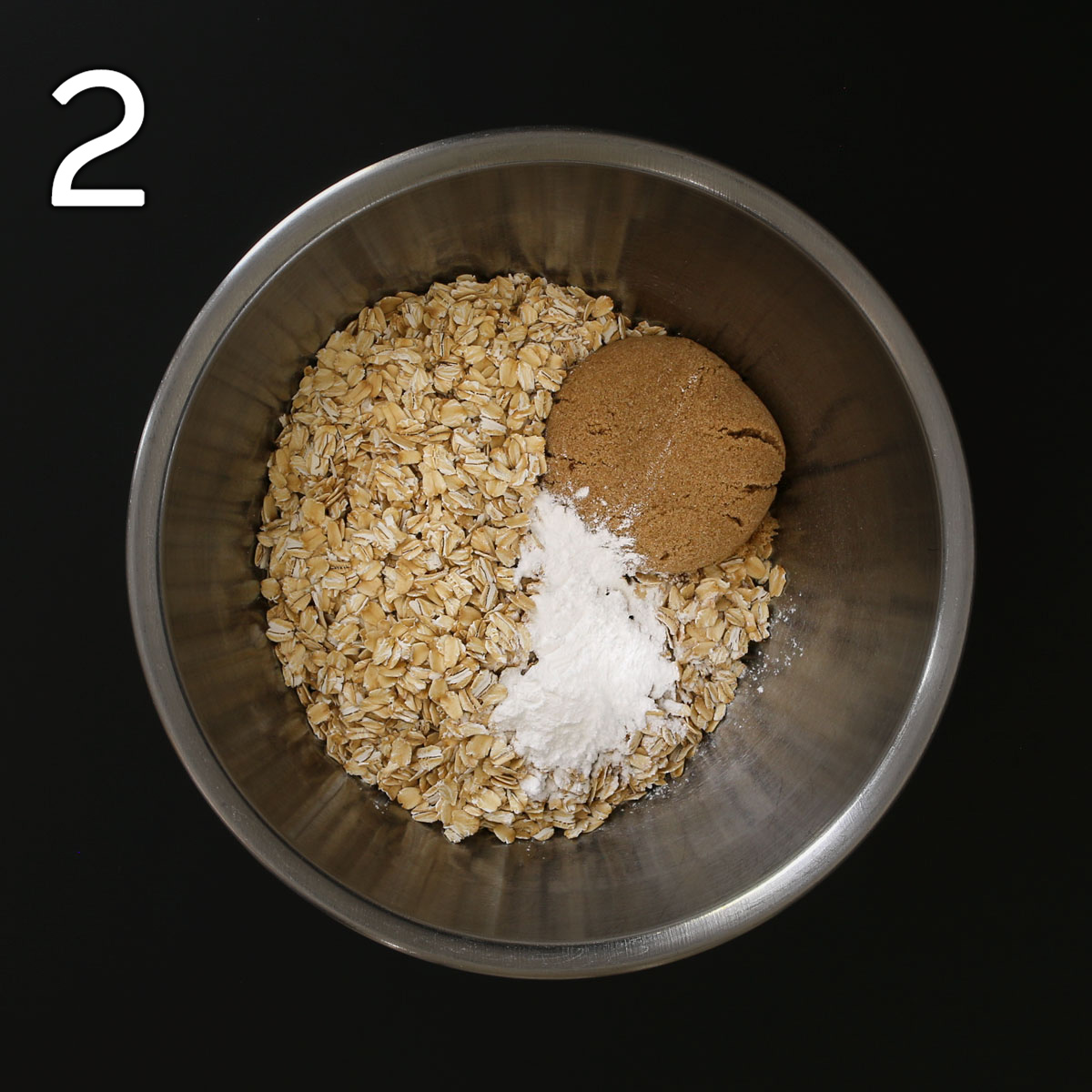 oats, leaveners, and brown sugar in large metal mixing bowl on black table top.