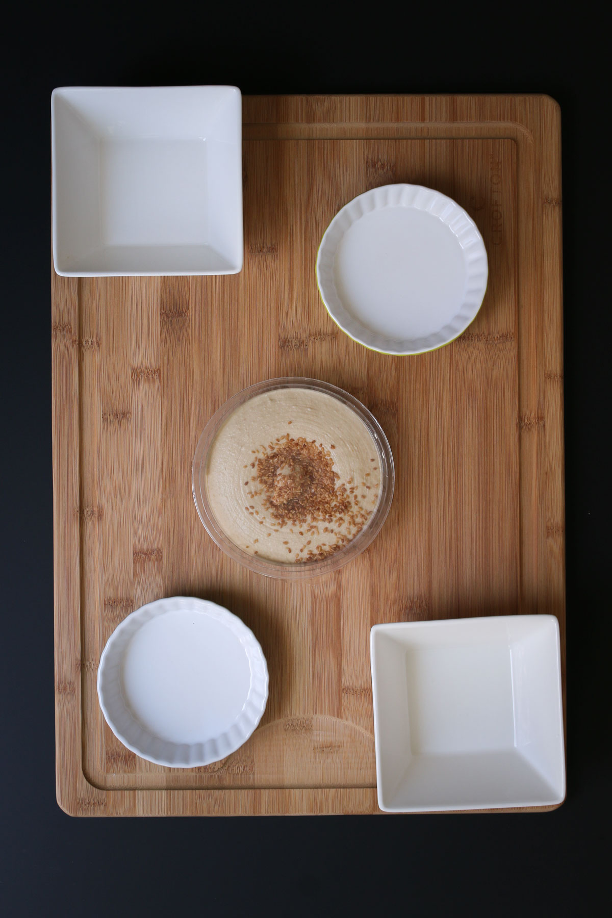 large wooden board with four white ceramic dishes spread out and a tub of hummus in the center.