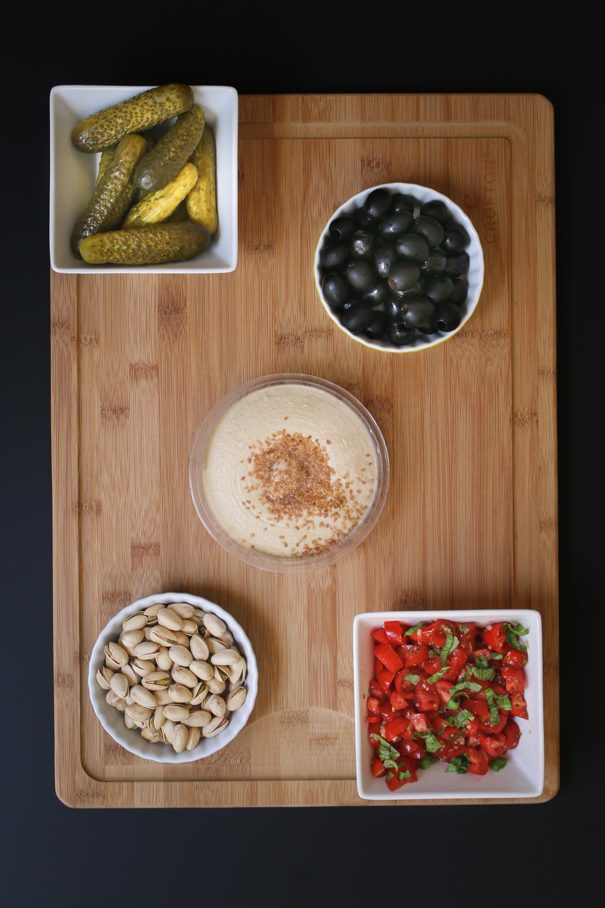 pickles and bruschetta in square bowls, nuts and olives in round ones on a wooden board with a tub of hummus.
