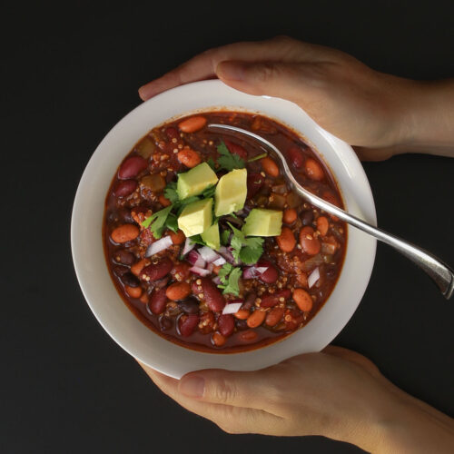 hands holding white bowl of quinoa chili with toppings on black table top.