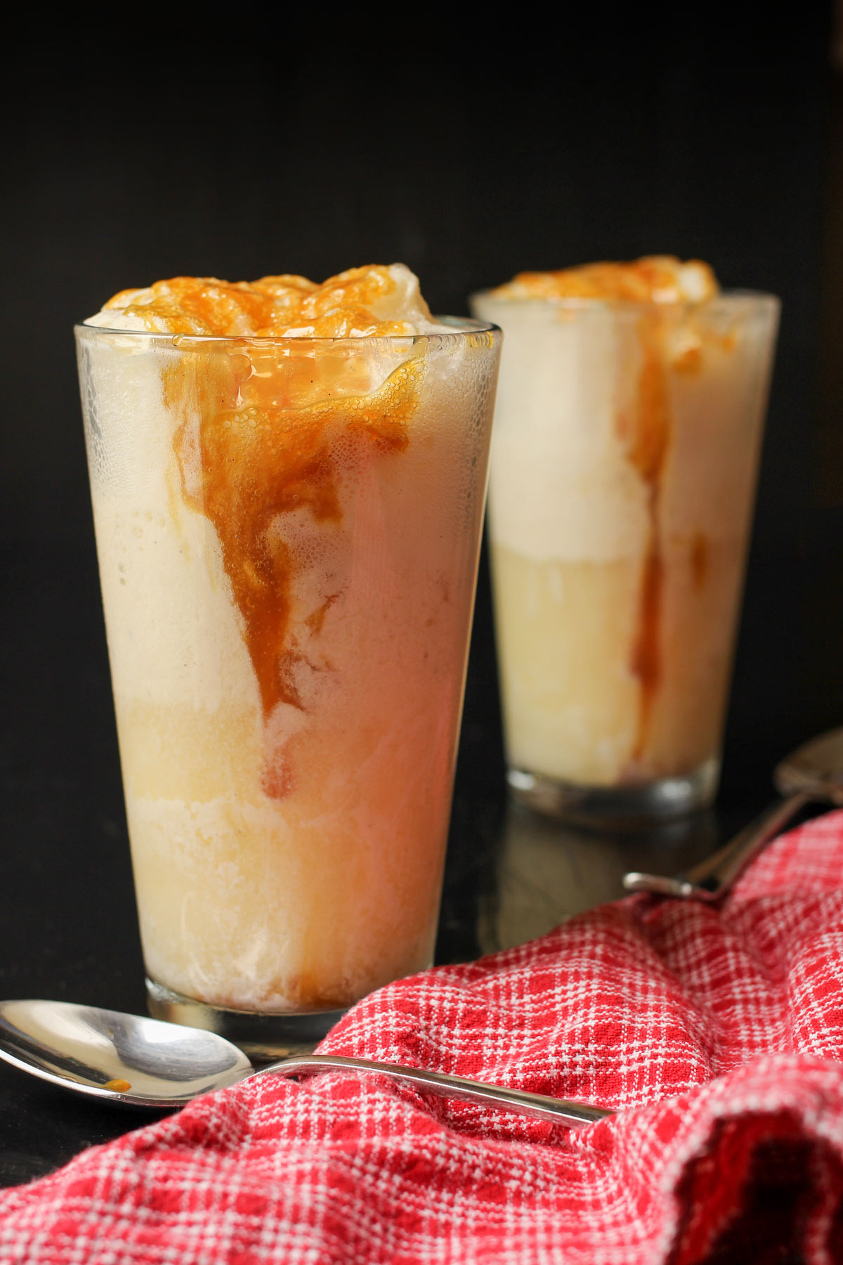 side view of two sparkling apple cider floats on a black table with a red plaid cloth nearby.