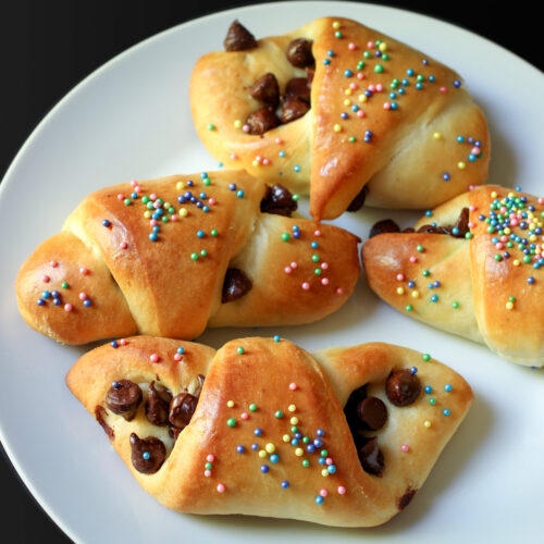 four chocolate butterhorn pastries with colored sprinkles on a white plate.