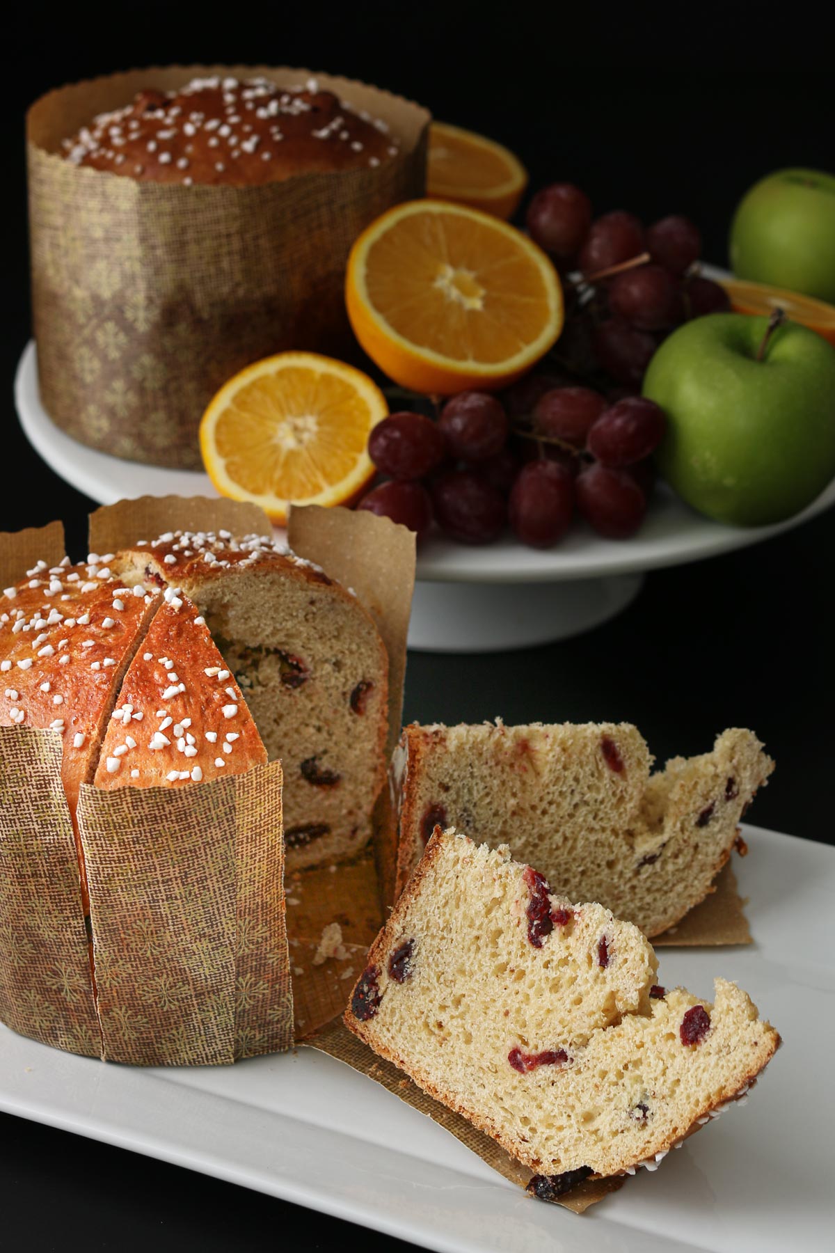 in the foreground a round of christmas bread sliced into wedges on a white platter, in the background a cake stand with another round of bread and assorted winter fruit.