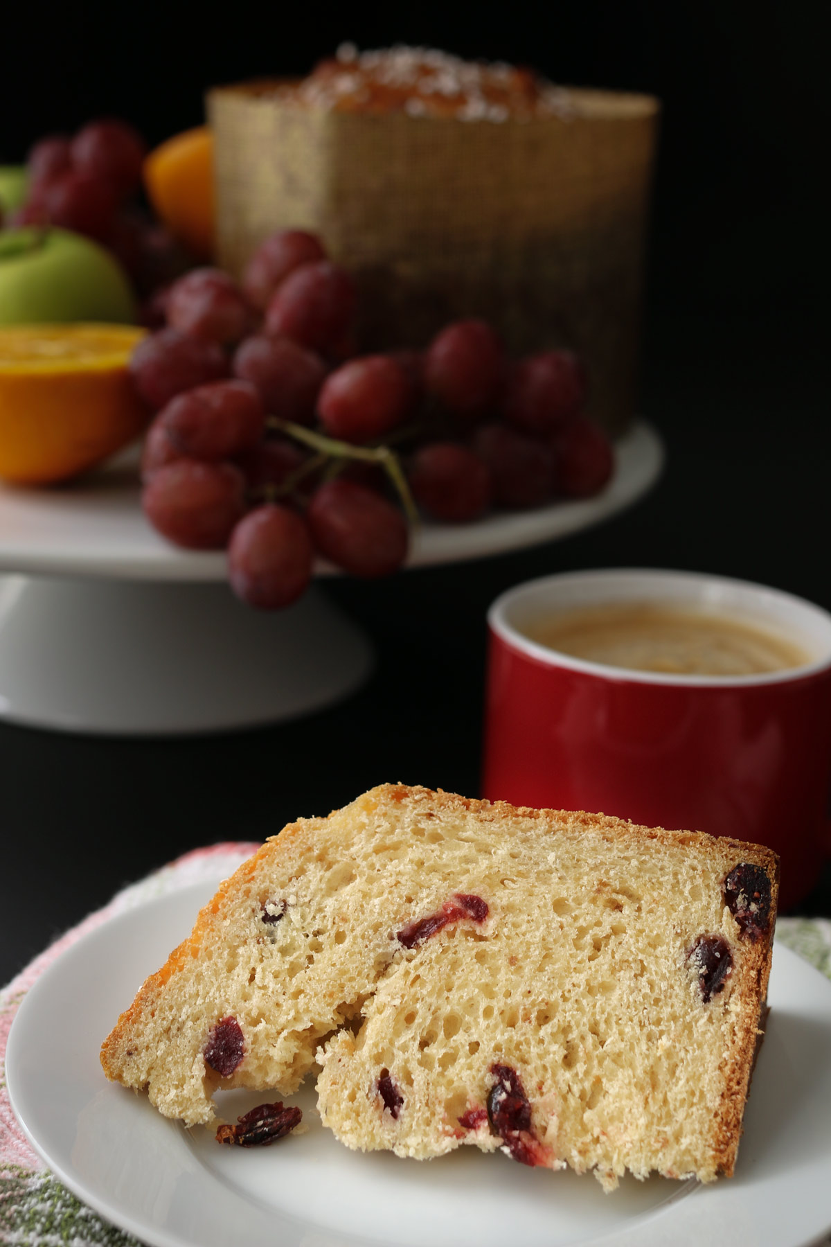 slice of cranberry christmas bread on a white plate with a cup of espresso and the cake stand of bread and fruit in the background.