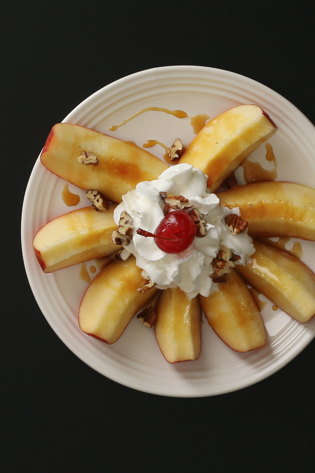 overhead shot of apple slices in a round with whipped cream, caramel, nuts, and a cherry on top, all on a rimmed white plate on a black table.