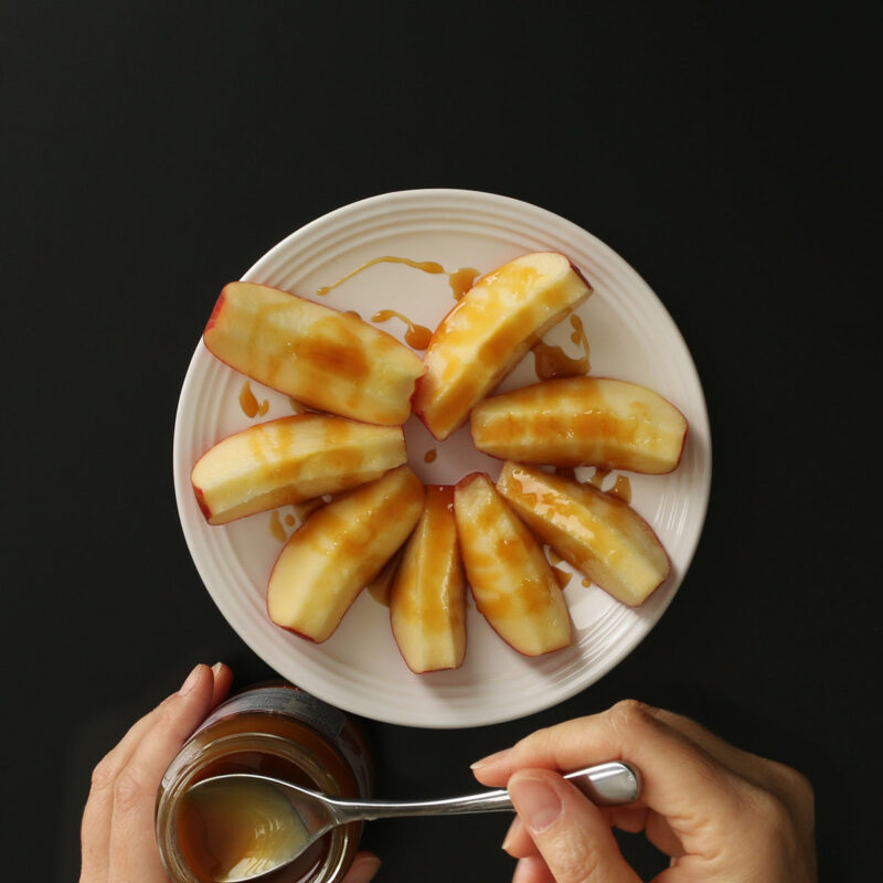dipping spoon into jar of caramel and drizzle over apple slices in a crown shape on a white plate.