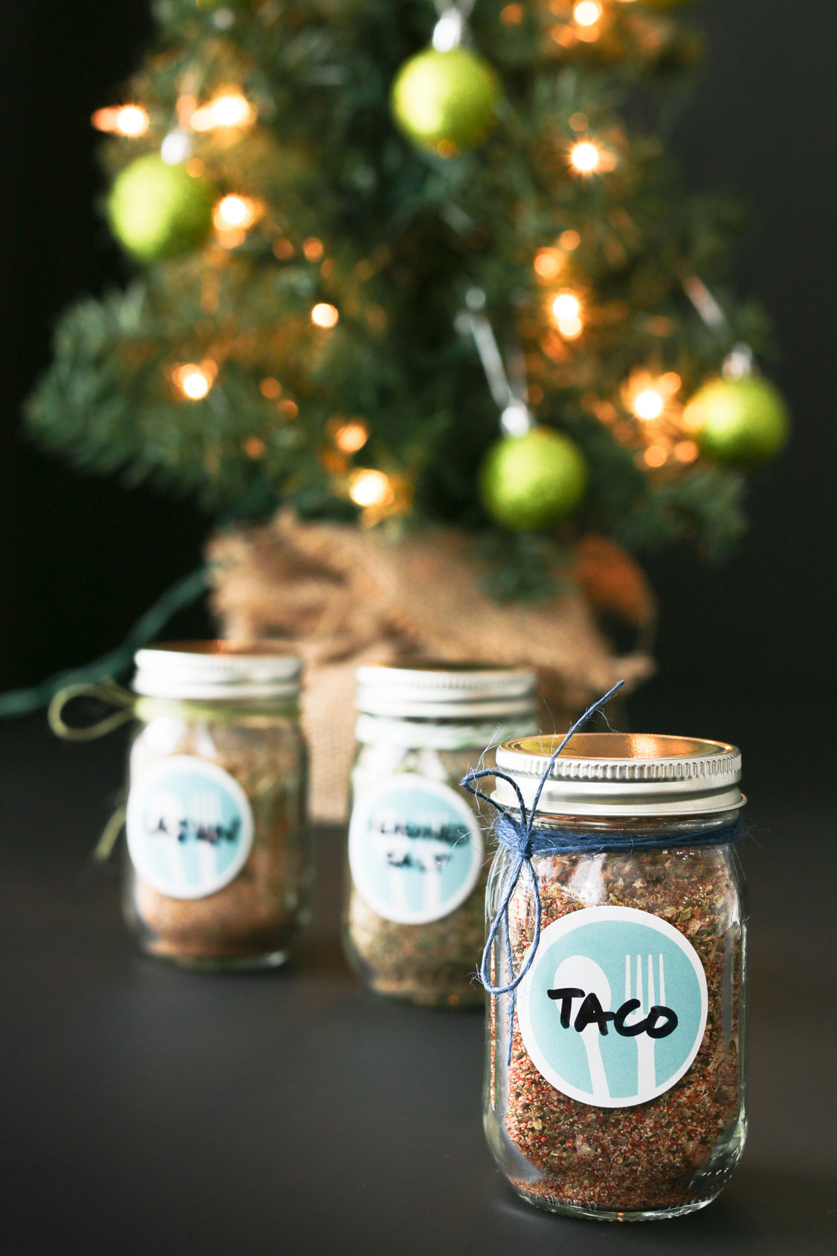 small jars of spice mixes on a black table near a lit christmas tree with green balls.