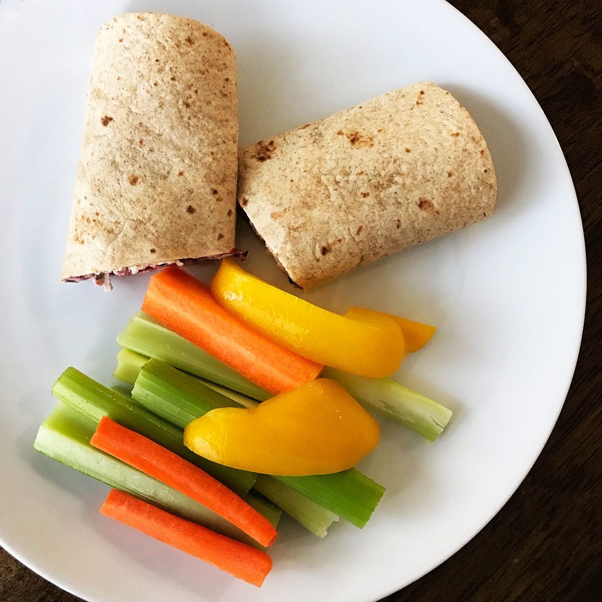 chicken salad wrap in whole wheat tortilla on white plate with crudite vegetables.