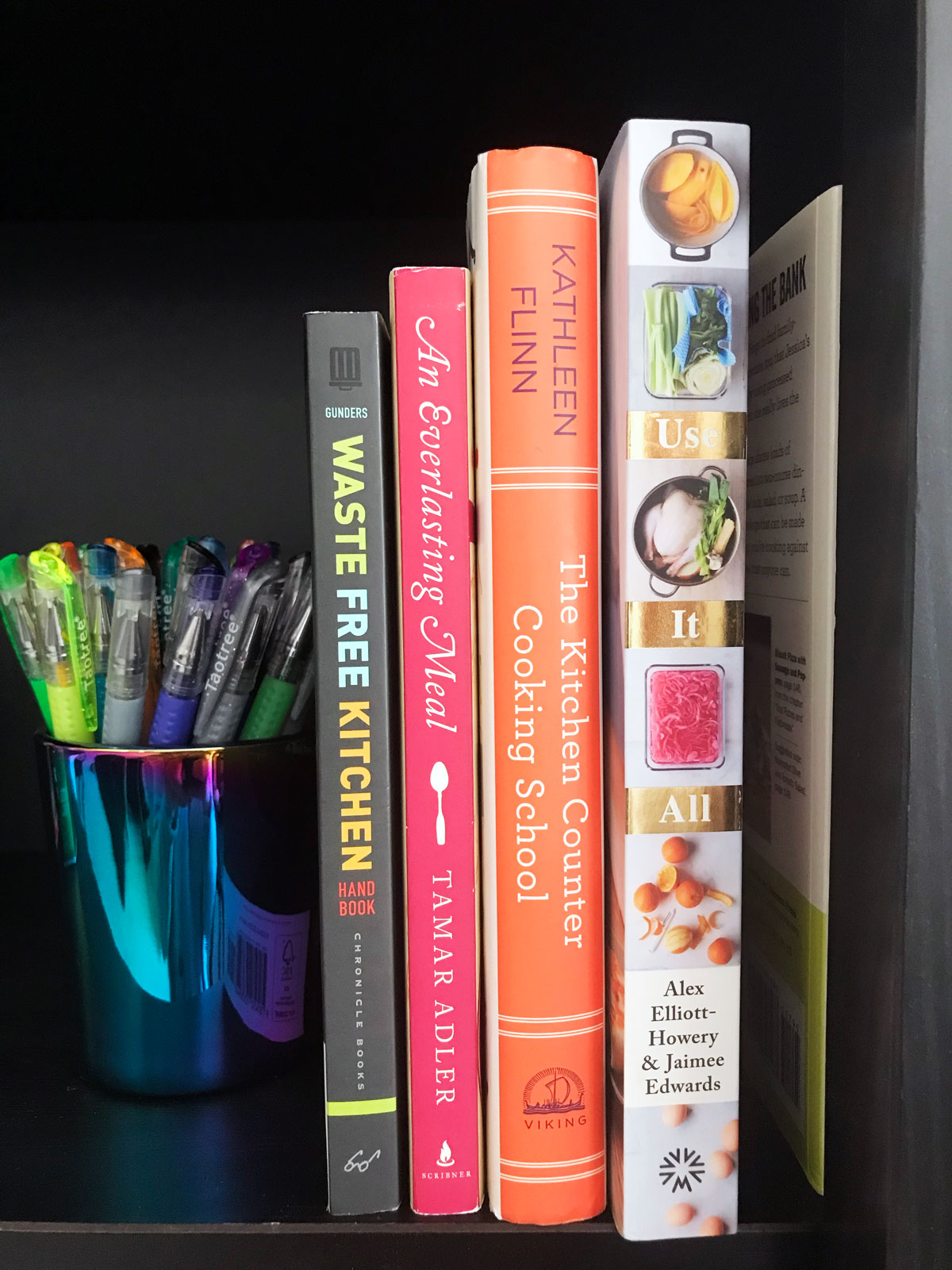 cookbooks lined up in black book case next to sparkly cup of gel pens.