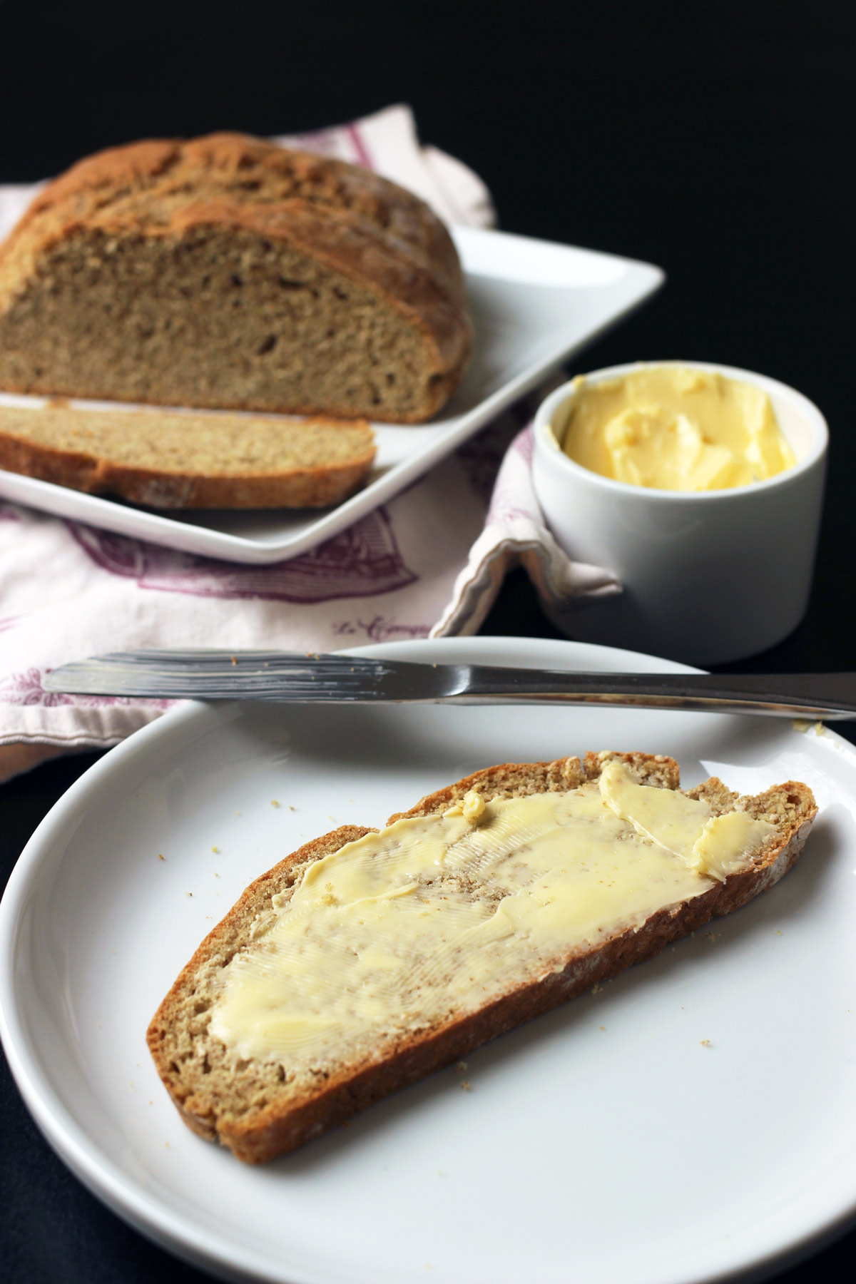 slice of irish soda bread on white plate slathered with butter with knife, butter dish, and loaf on plate nearby.