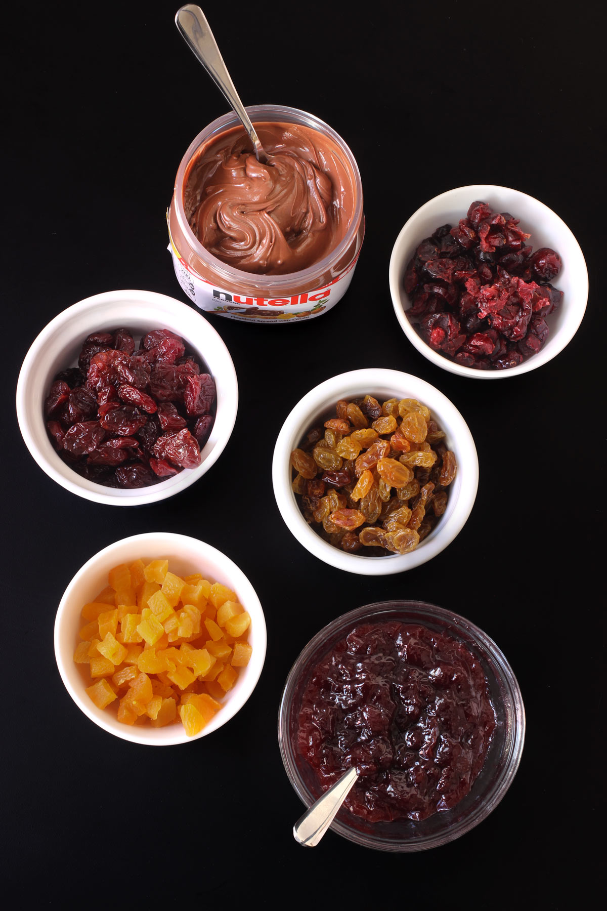 different types of dried fruit and spreads in white dishes laid out on a black table.