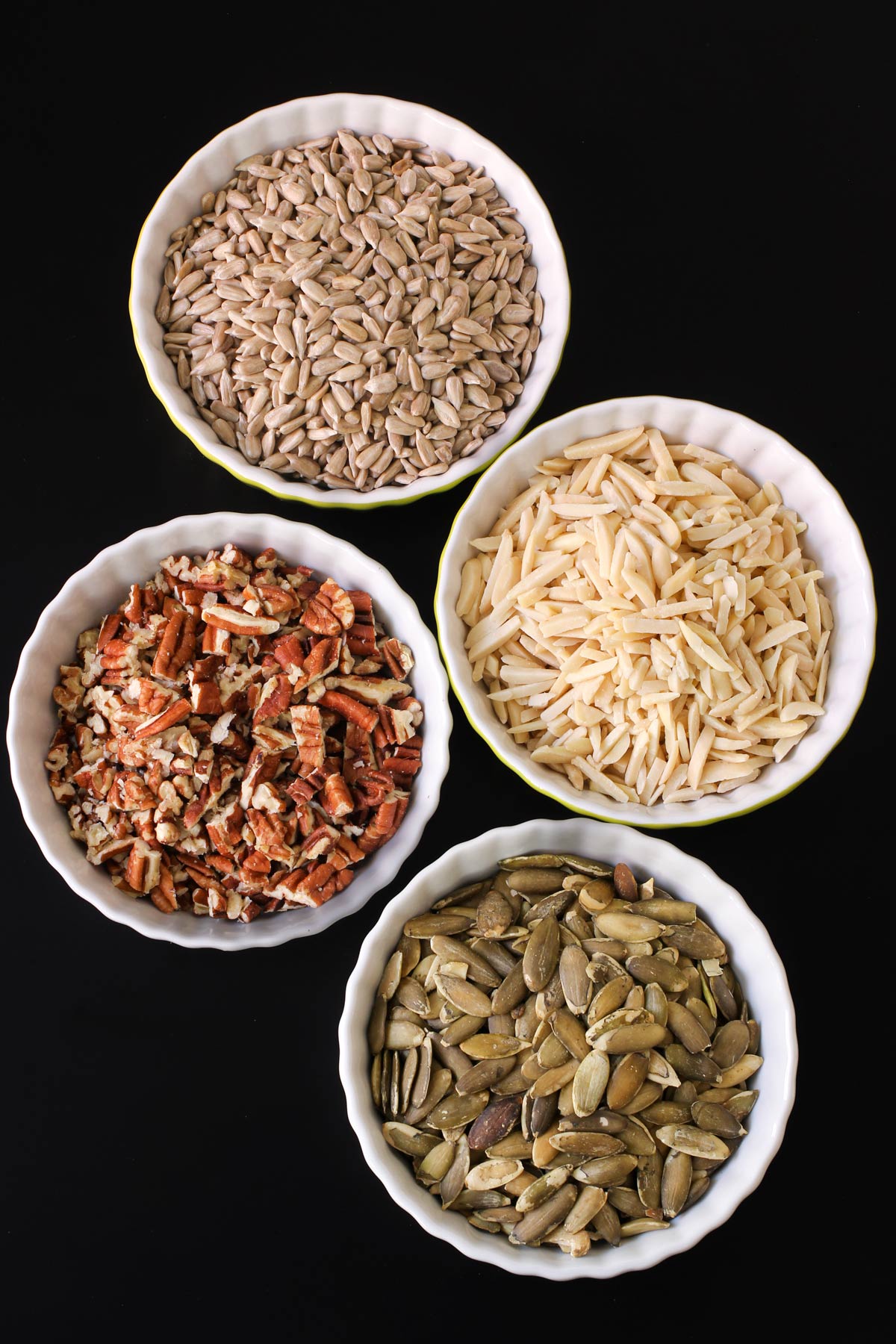 different types of nuts and seeds in white dishes on a black table top.