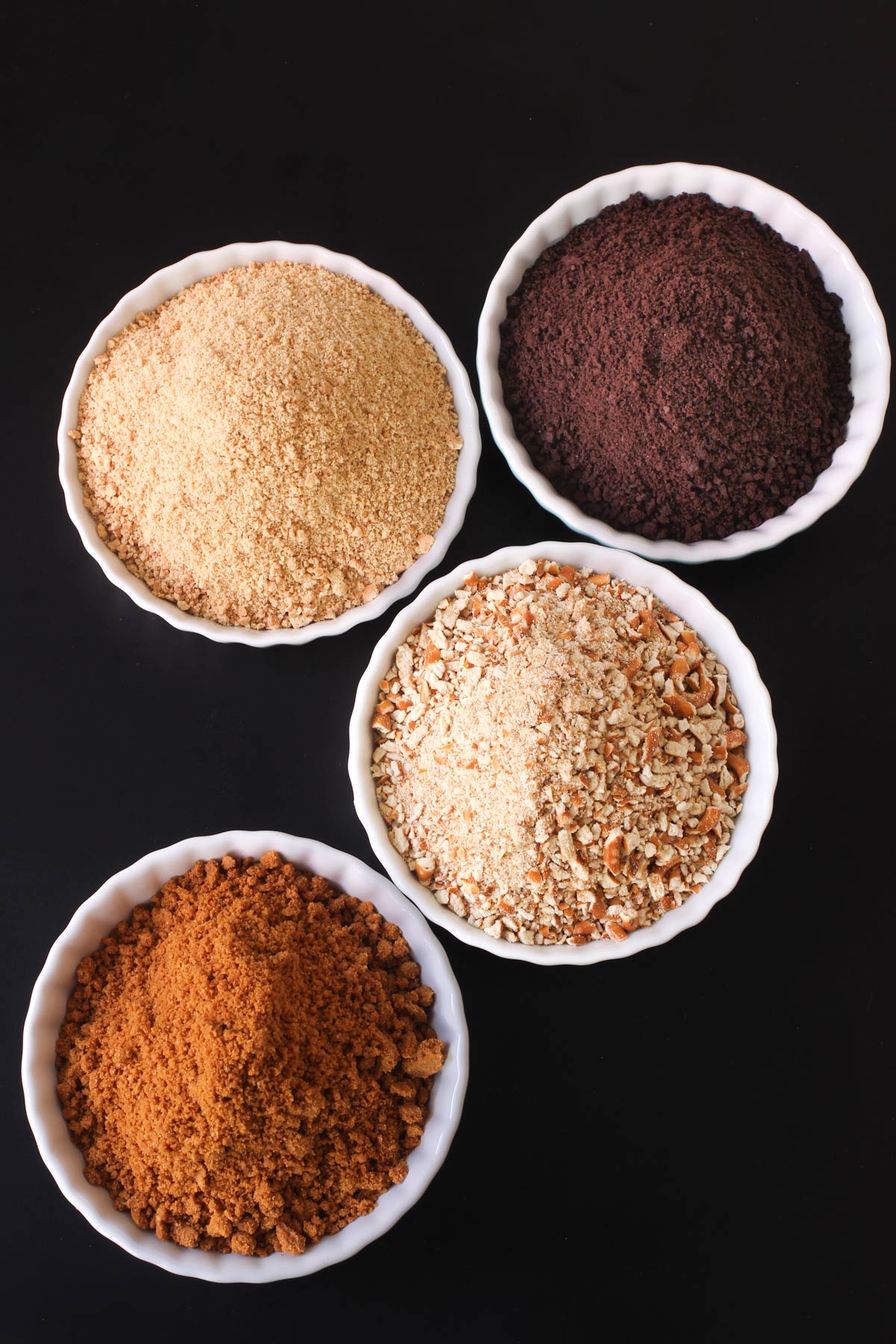 different cracker and cookie crumbs in small white dishes on black table.