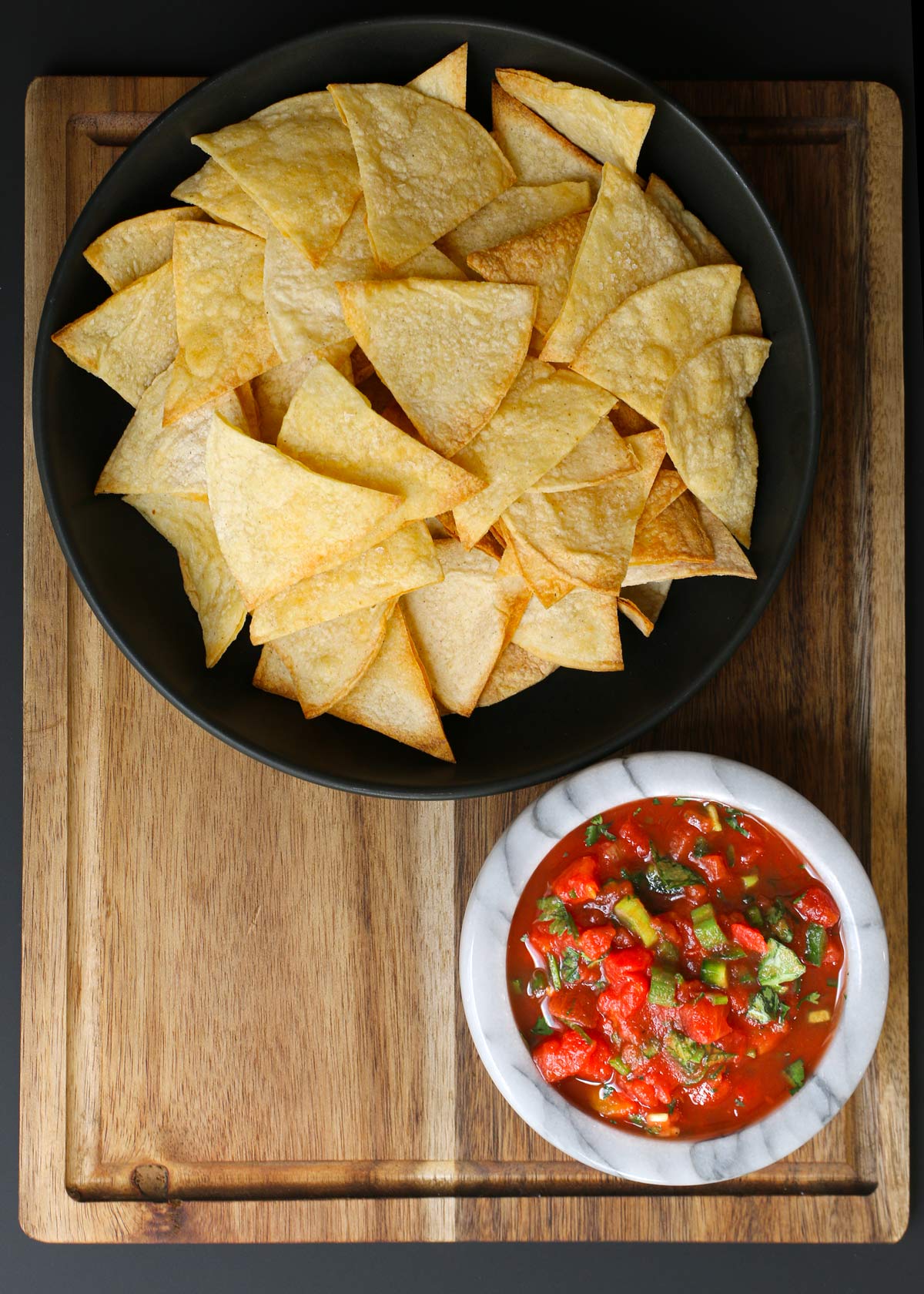 black bowl of tortilla chips and marble bowl of salsa on a wood board against a black backdrop.