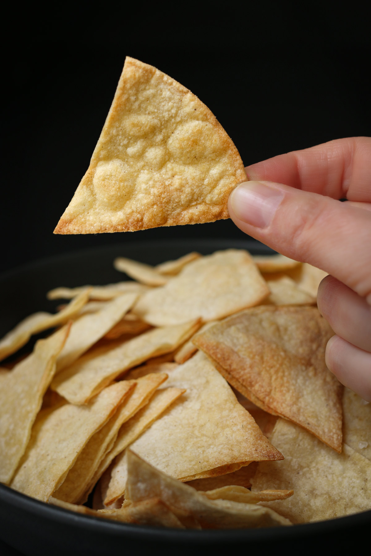 hand holding a triangle tortilla chip over a black plate of air fryer tortilla chips with a black background.