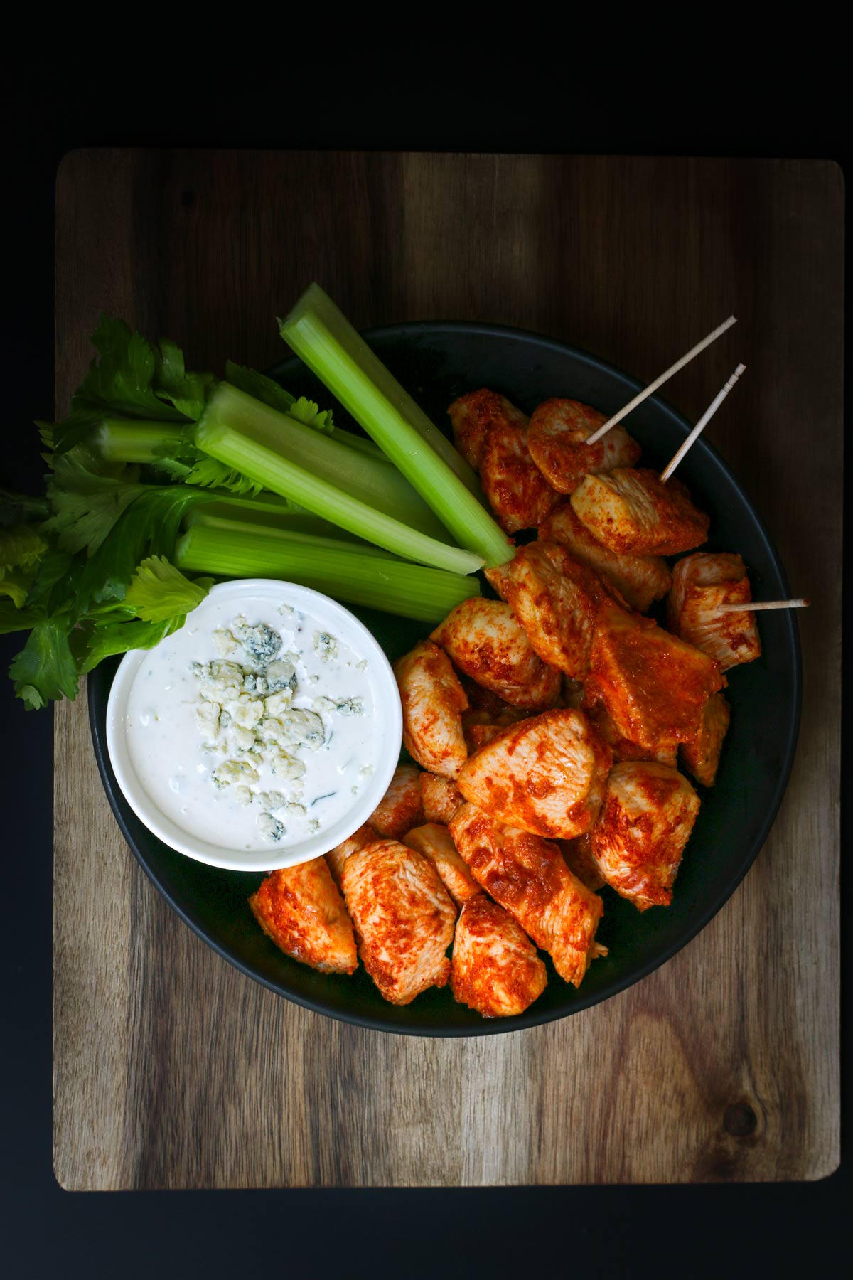 overhead shot of black plate on wooden board with buffalo chicken bites, some with toothpicks, an array of celery sticks, and a small white bowl of blue cheese dressing.