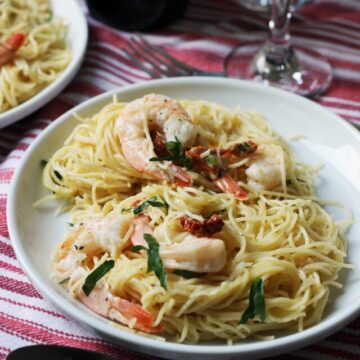 close up of a plate of shrimp pasta next to wine glass on red and white striped cloth.