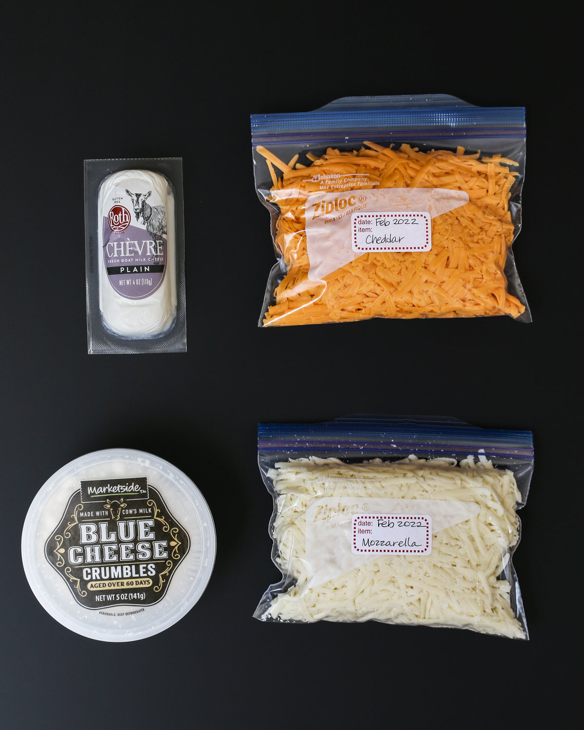package of goat cheese, tub of blue cheese crumbles, and bags of cheddar and mozzarella cheese on black table.