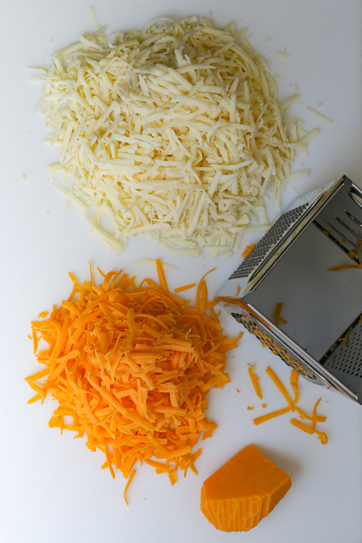 piles of jack and cheddar cheese on white board next to box grater with a small hunk of cheddar nearby.