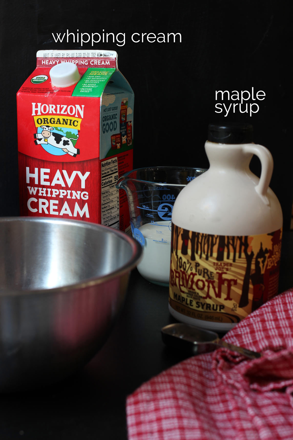 carton of whipped cream near a bottle of maple syrup on black table with bowl and measure and red cloth.