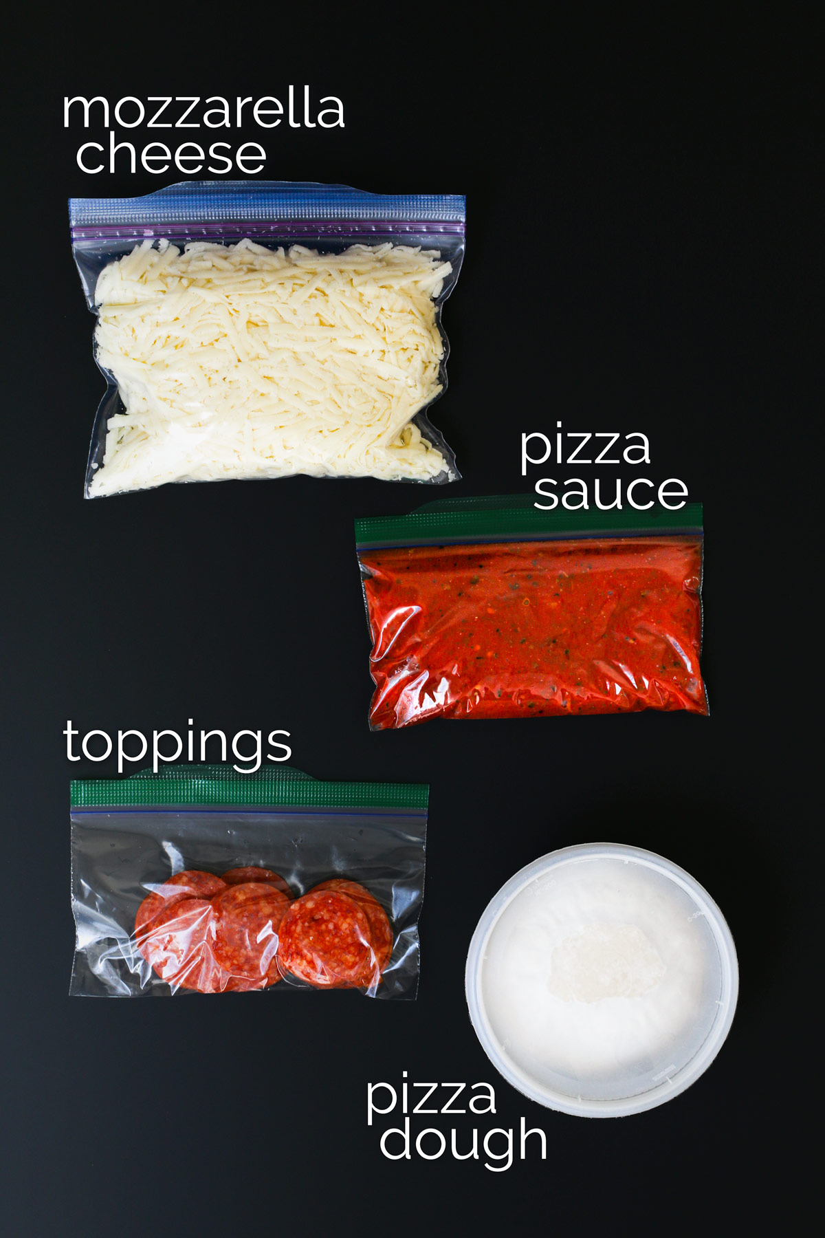 ingredients for pizza kits packaged for freezing spread out on black table.