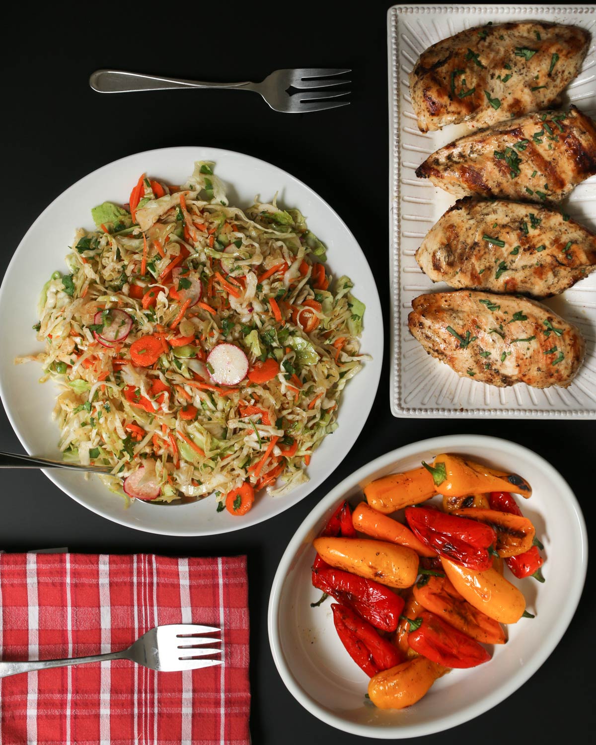 dinner table set with bowl of Mexican coleslaw, platter of grilled chicken, and platter of grilled mini peppers.