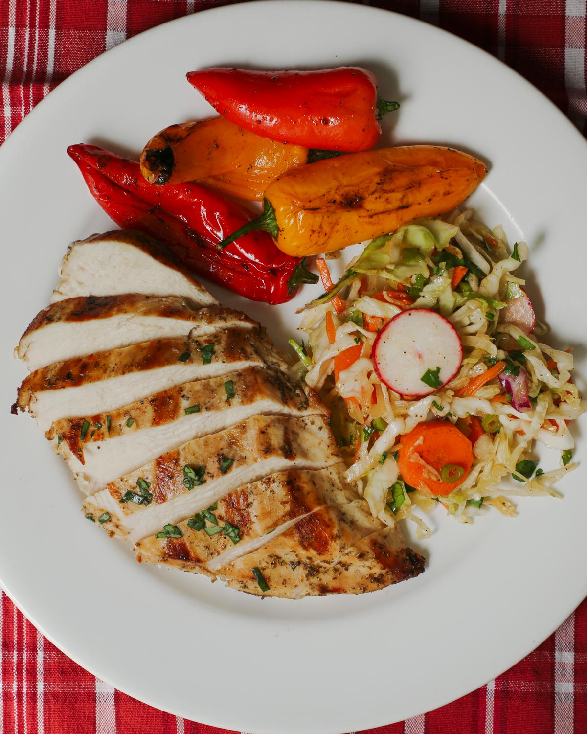 white dinner plate with grilled peppers, coleslaw, and sliced chicken breast on a red plaid cloth.