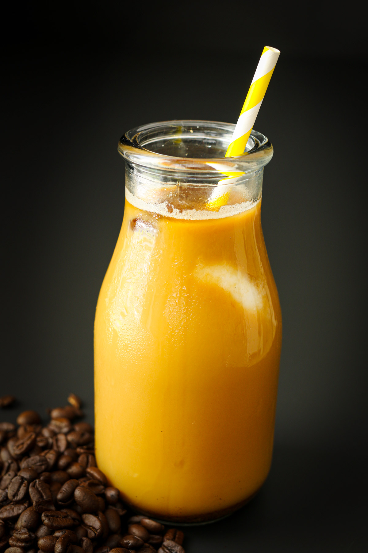 iced coffee in a glass milk bottle with coffee beans surrounding its base on a black table.