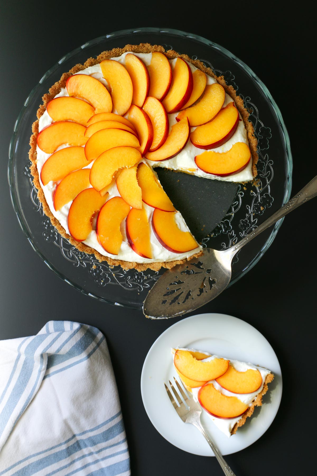 nectarine pie on glass serving platter with a slice cut out on a plate nearby with blue and white napkin on the table.