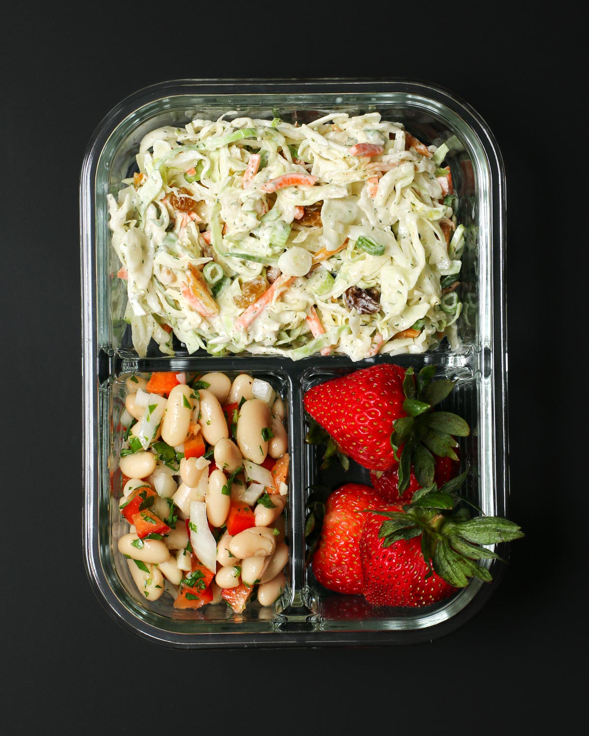 divided glass meal prep box with coleslaw, bean salad, and strawberries in the sections.