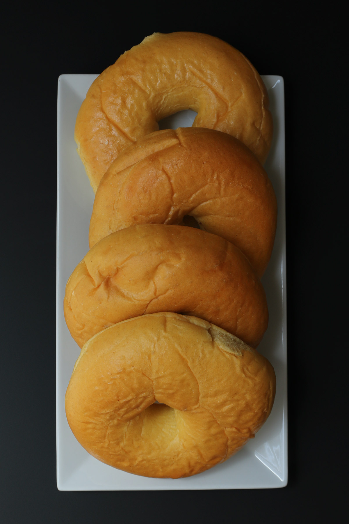 four bagels on a rectangular, white platter on a black table.