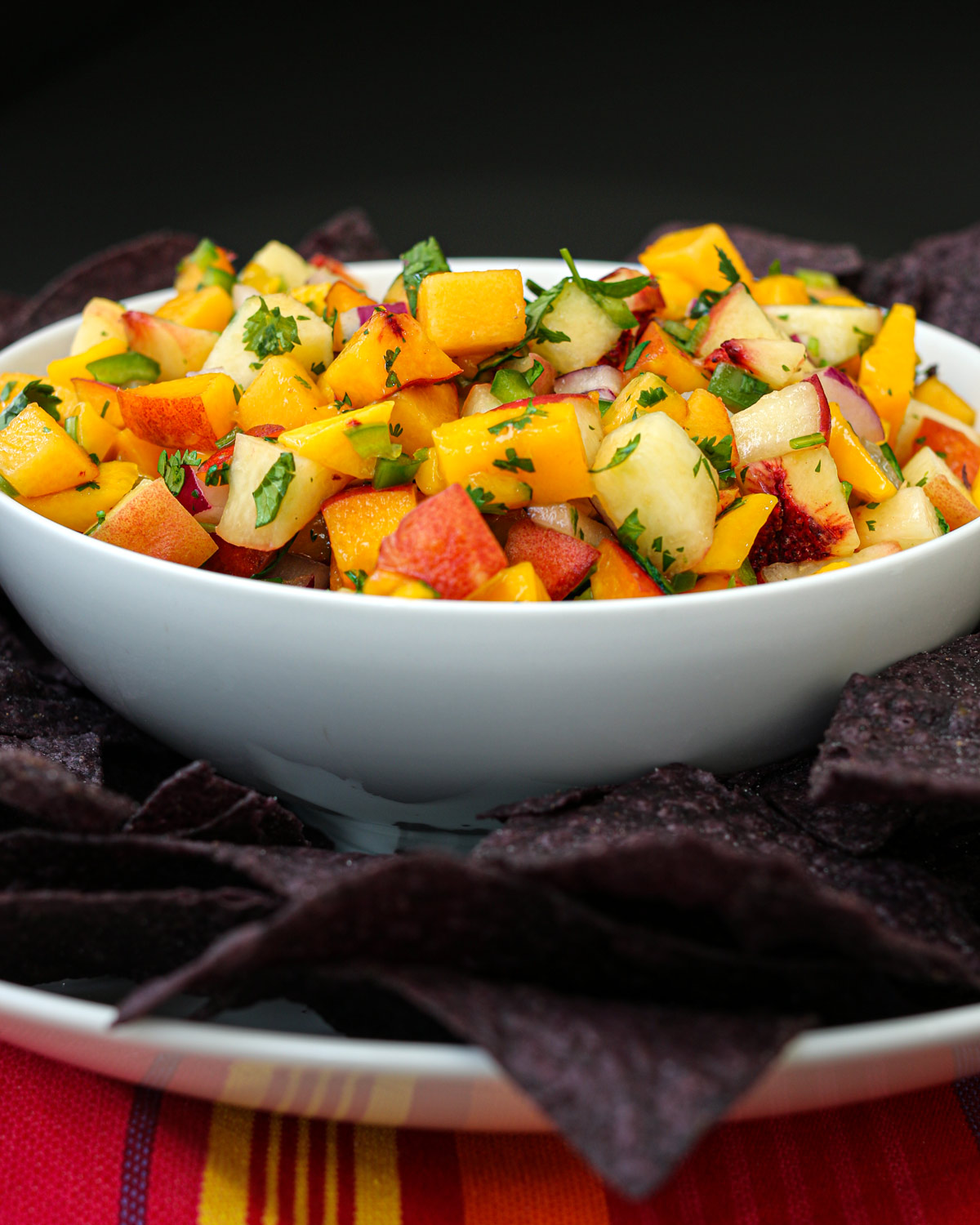bowl of peach salsa surrounded by blue corn tortilla chips on a platter.