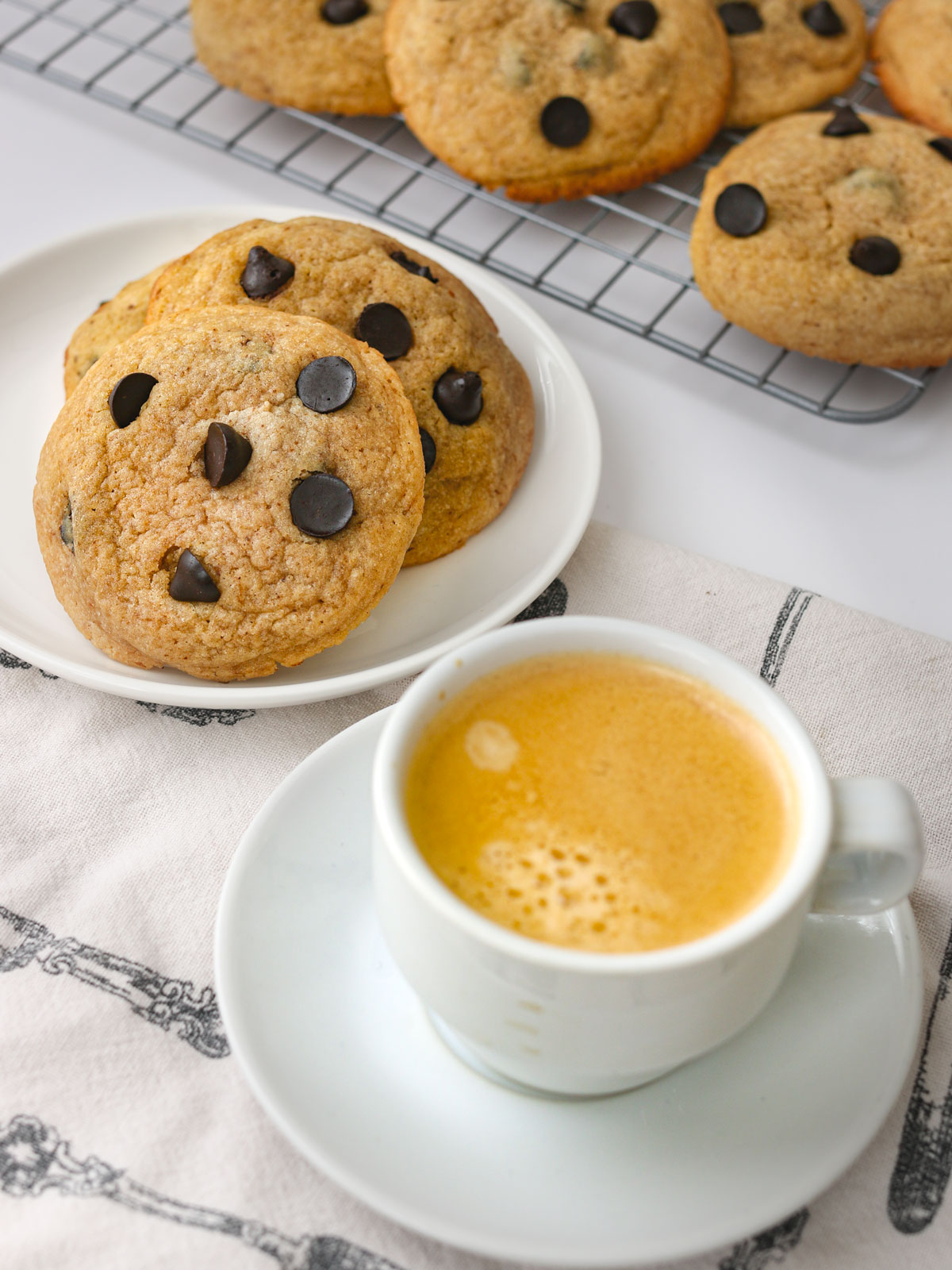 espresso cup and saucer next to plate of cookies with tray of cookies in background.