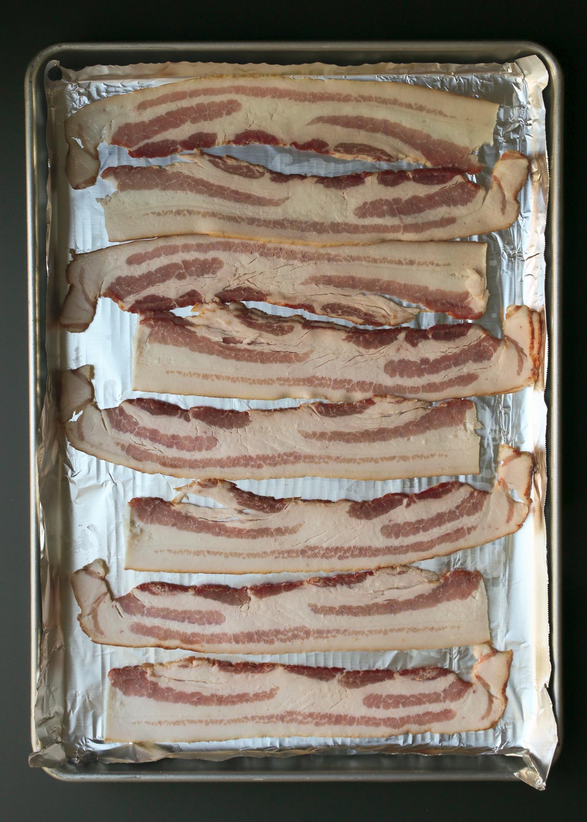 uncooked bacon laid out on rimmed baking sheet.