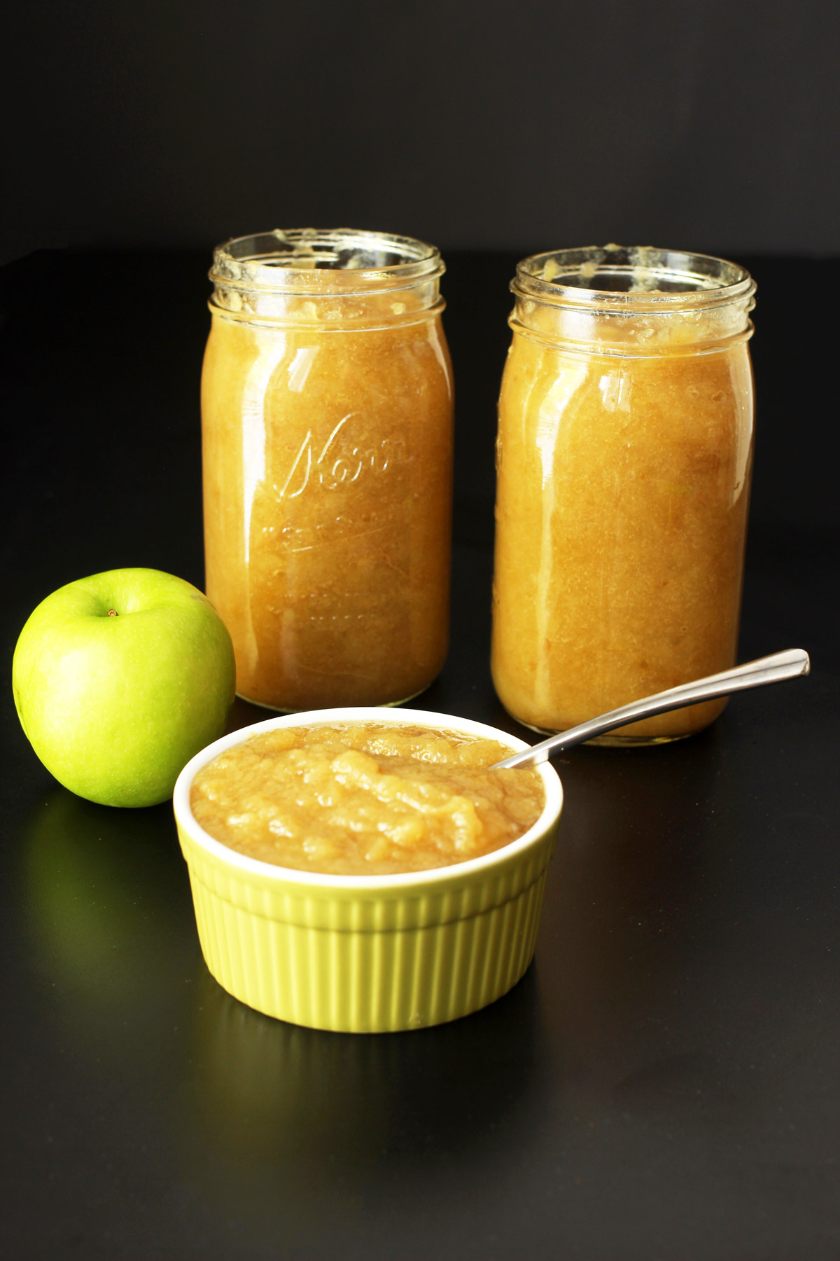 two jars of applesauce next to an apple and a ramekin of sauce.