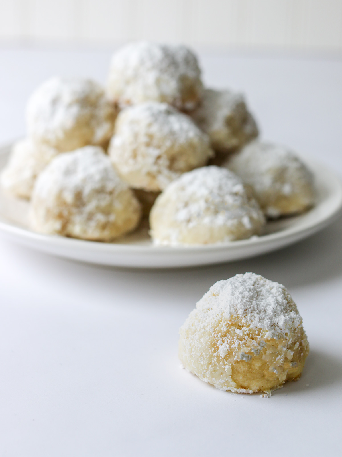 a snowball cookie on a white surface next to a white plate of stacked cookies.