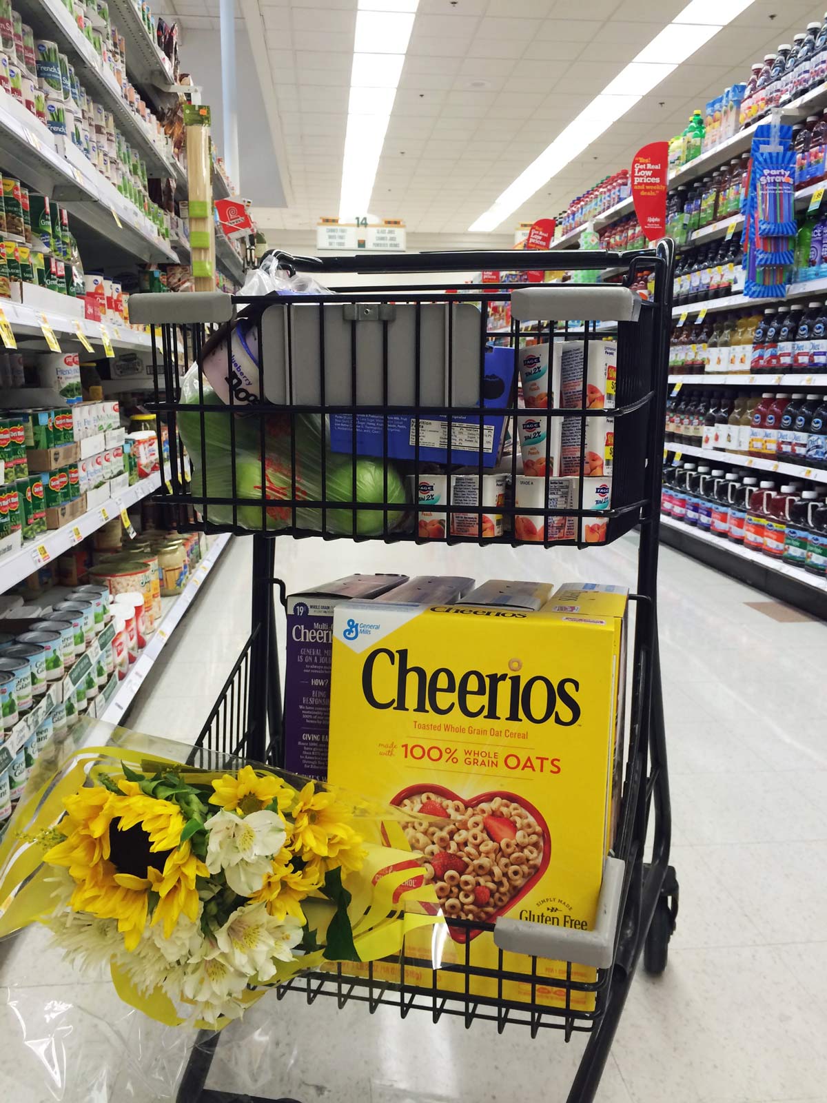 close up of shopping cart with flowers and Cheerios in the bottom basket, in grocery aisle.