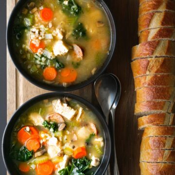 two bowls of turkey rice soup on board with spoons and sliced baguette.