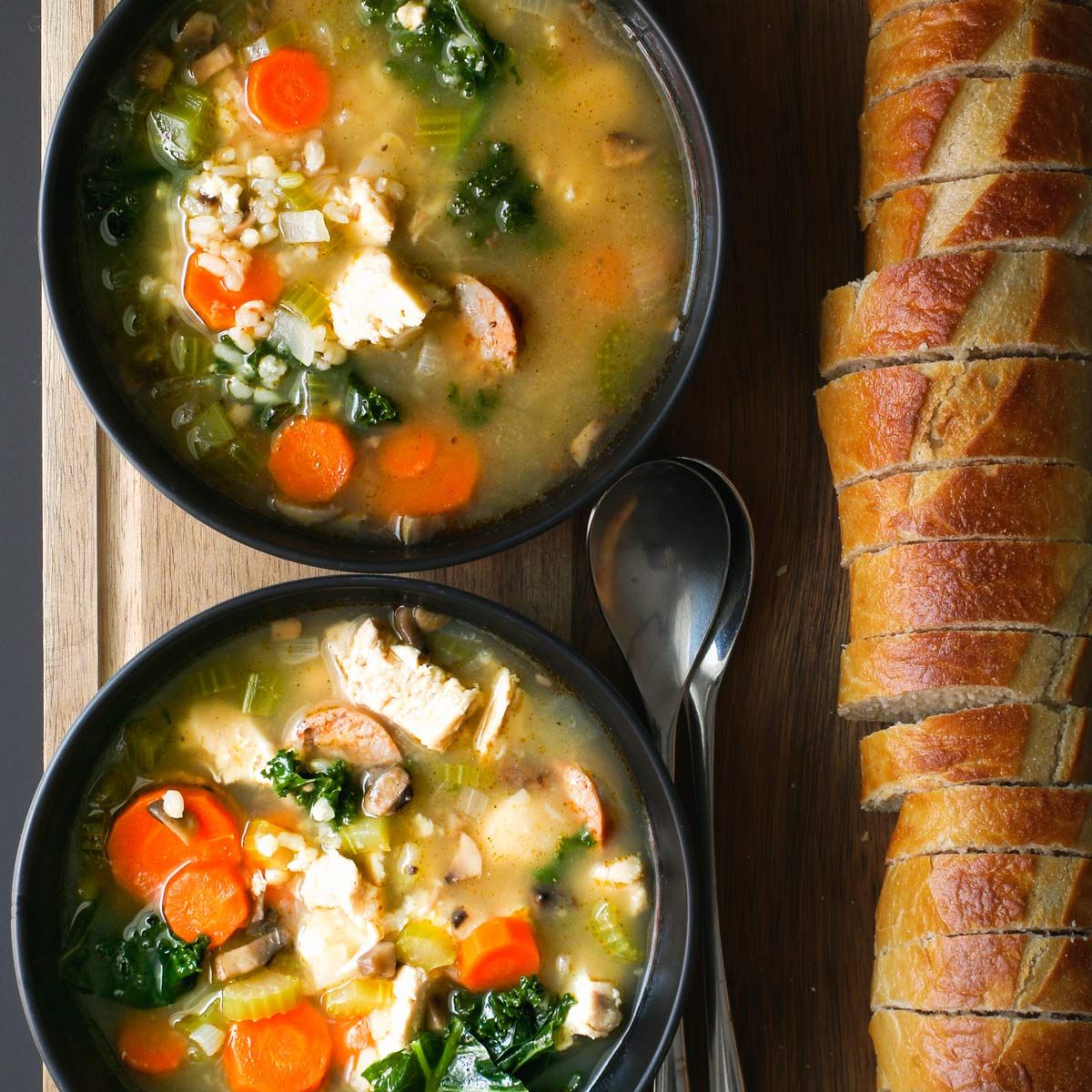 two bowls of turkey rice soup on board with spoons and sliced baguette.