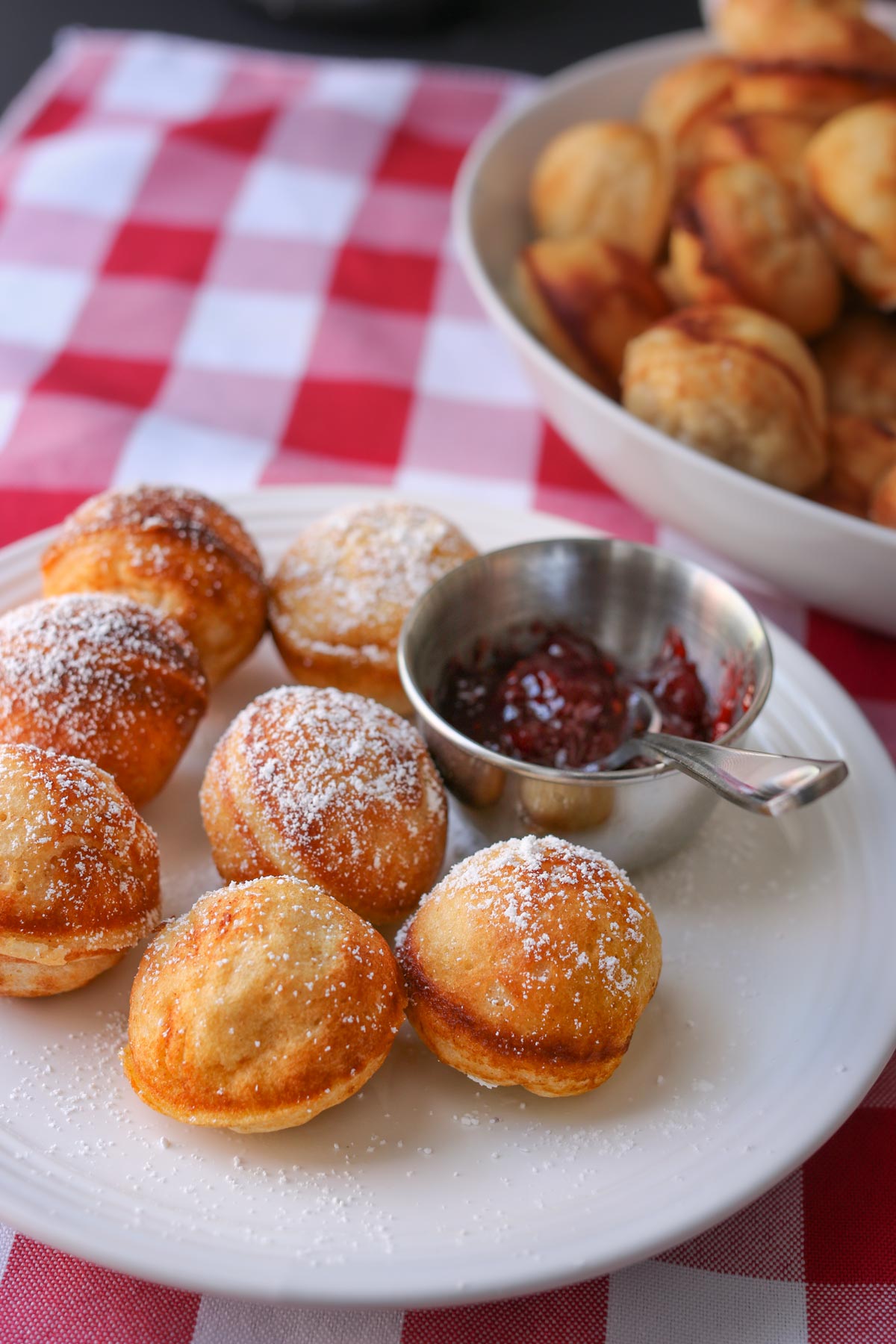 plate of aebleskiver sprinkled with powdered sugar, alongside small metal cup of raspberry jam with a tasting spoon.