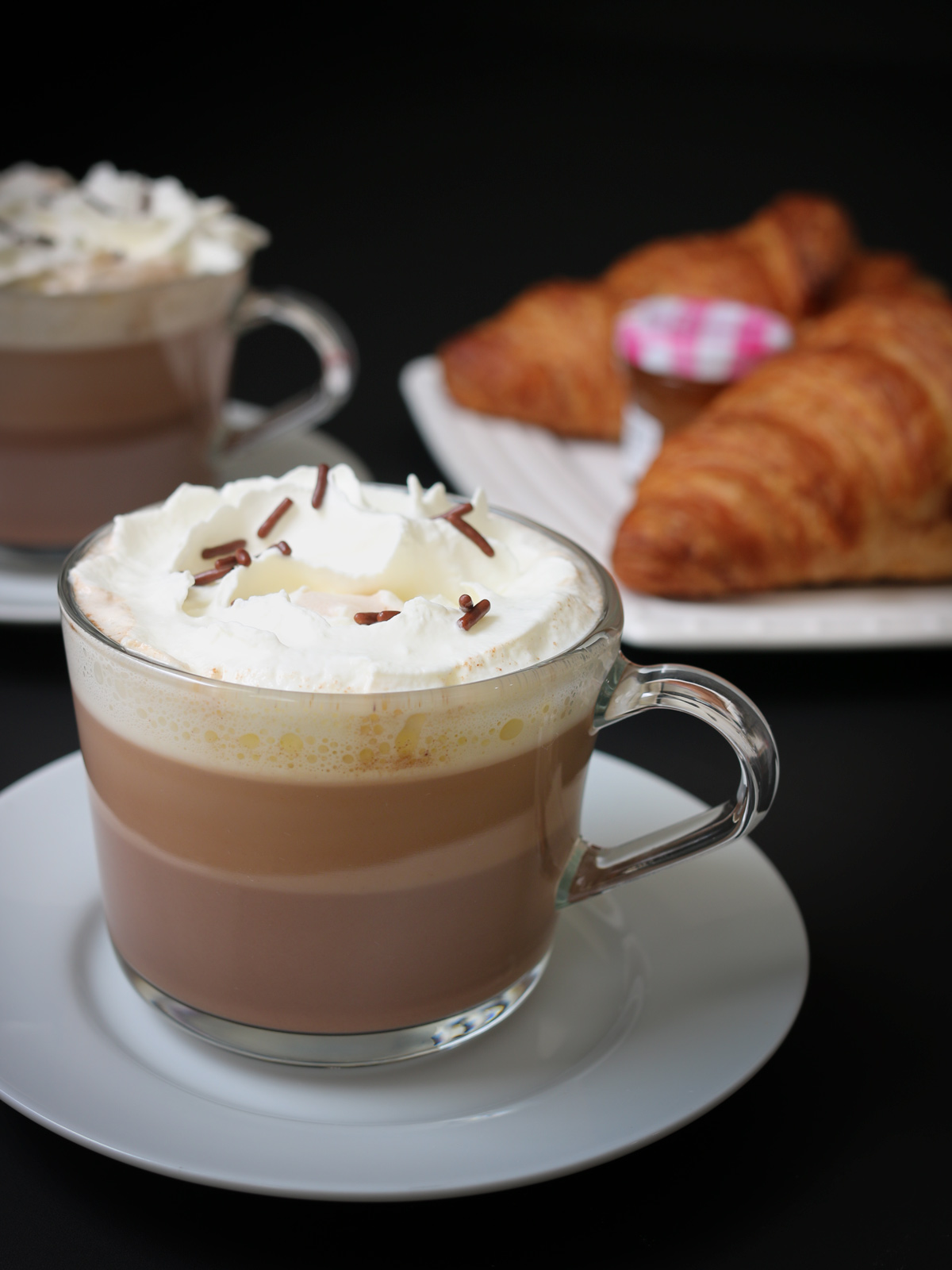 clear mugs of hot chocolate with toppings on white saucers, a plate of croissant in the background.