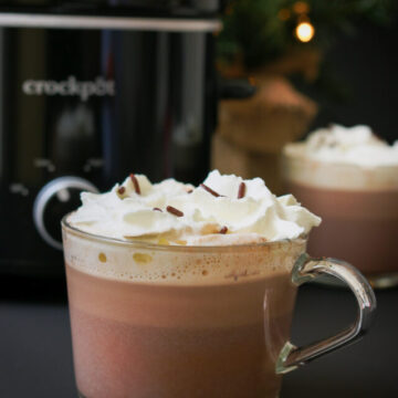 glass mugs of hot chocolate in front of small tabletop Christmas tree and a slow cooker.