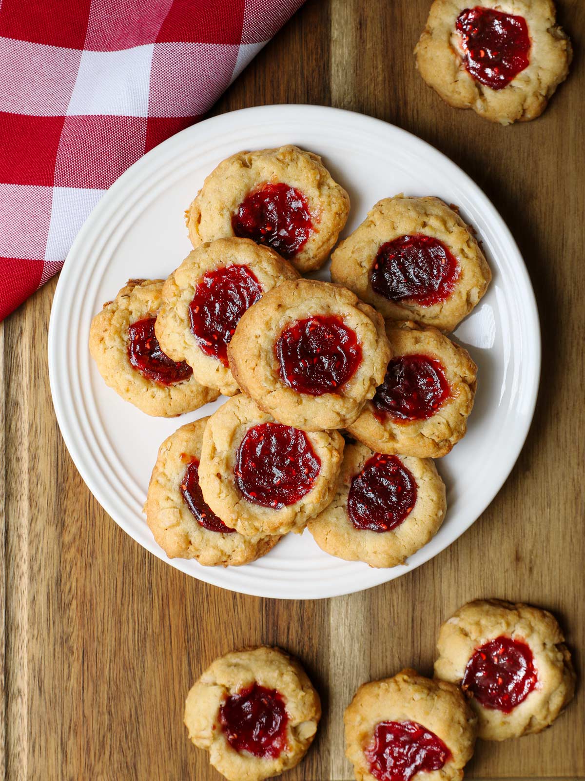 plate of raspberry thumbprint cookies on a wood board with more cookies and a red checked cloth.