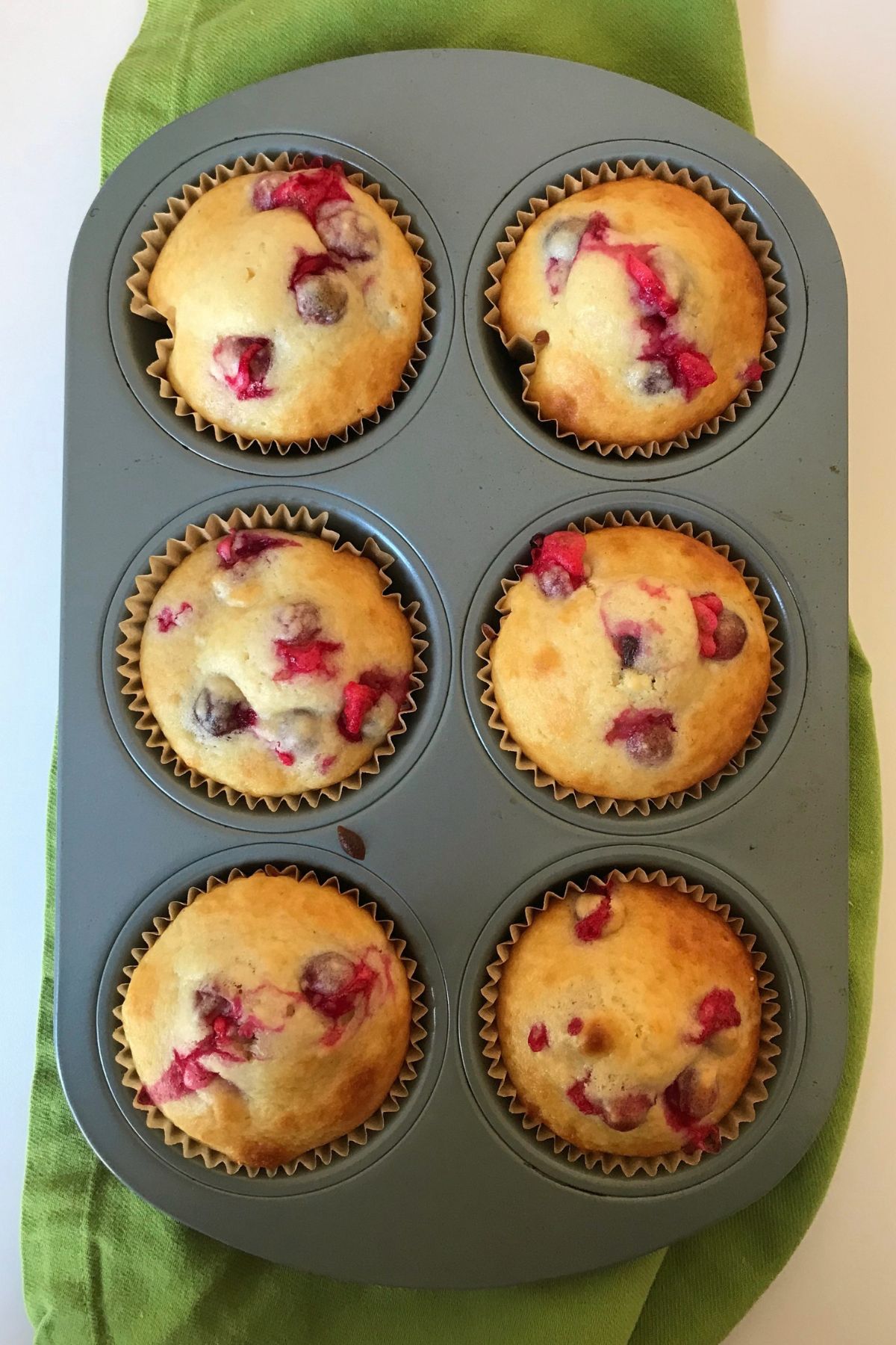 six baked cranberry muffins in muffin tin atop green cloth.