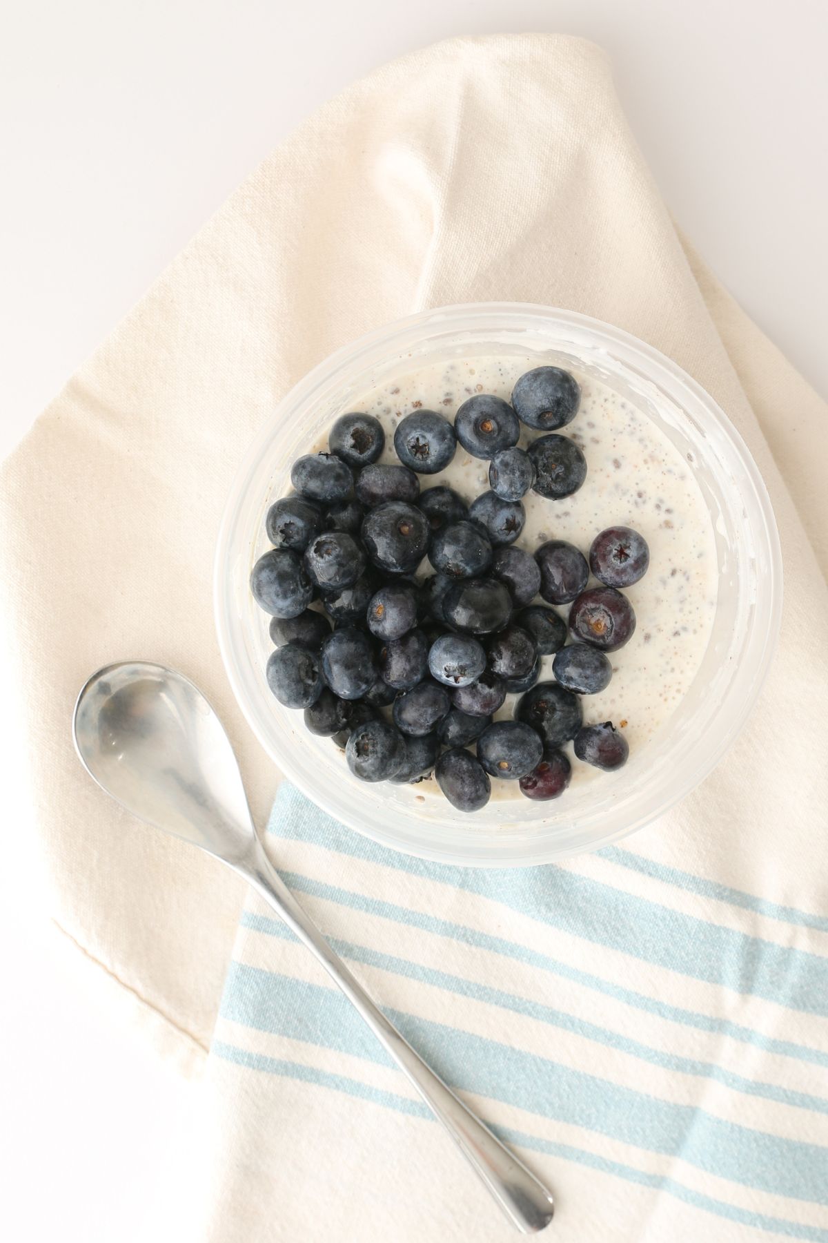 overnight protein oats in clear container topped with blueberries, with a tea towel and spoon.