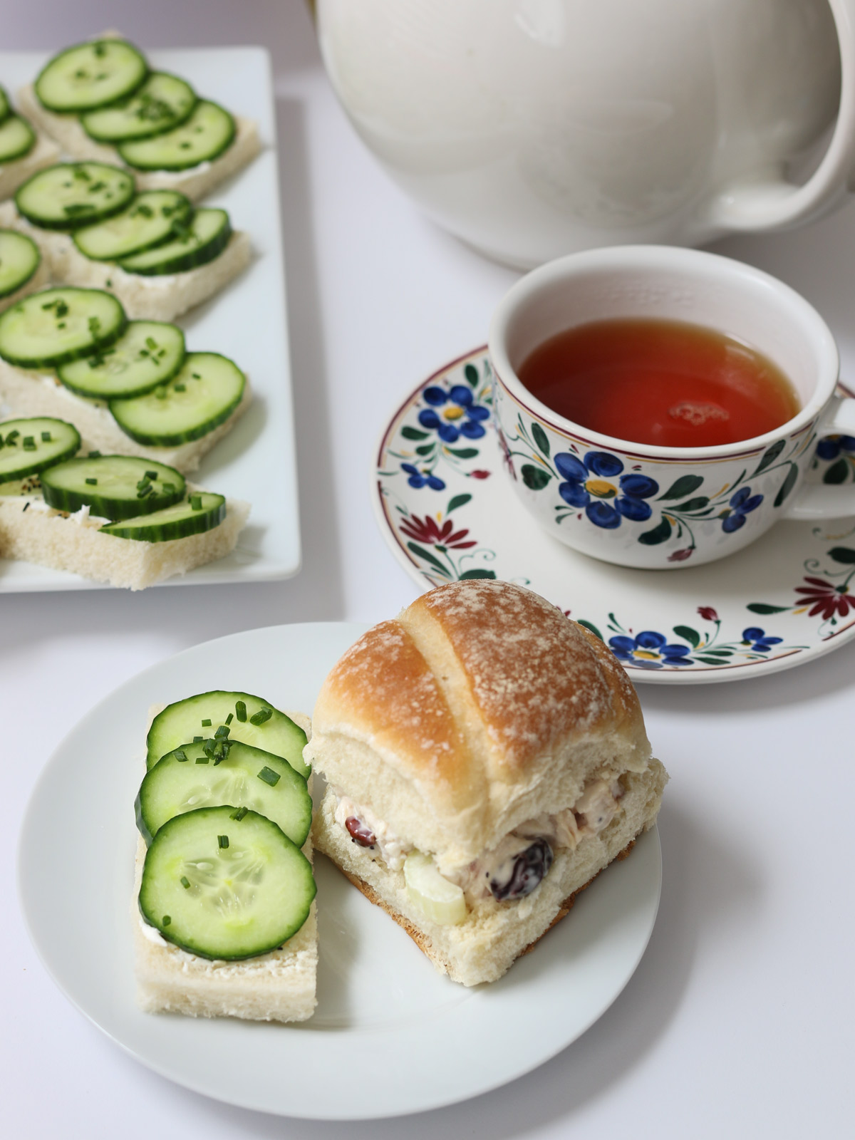 cucumber sandwich on plate with a small chicken salad sandwich, platter and tea behind them on table.
