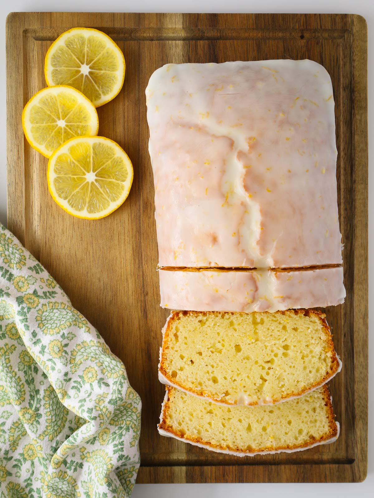lemon loaf on board, sliced, with lemon slices and a yellow and green towel.